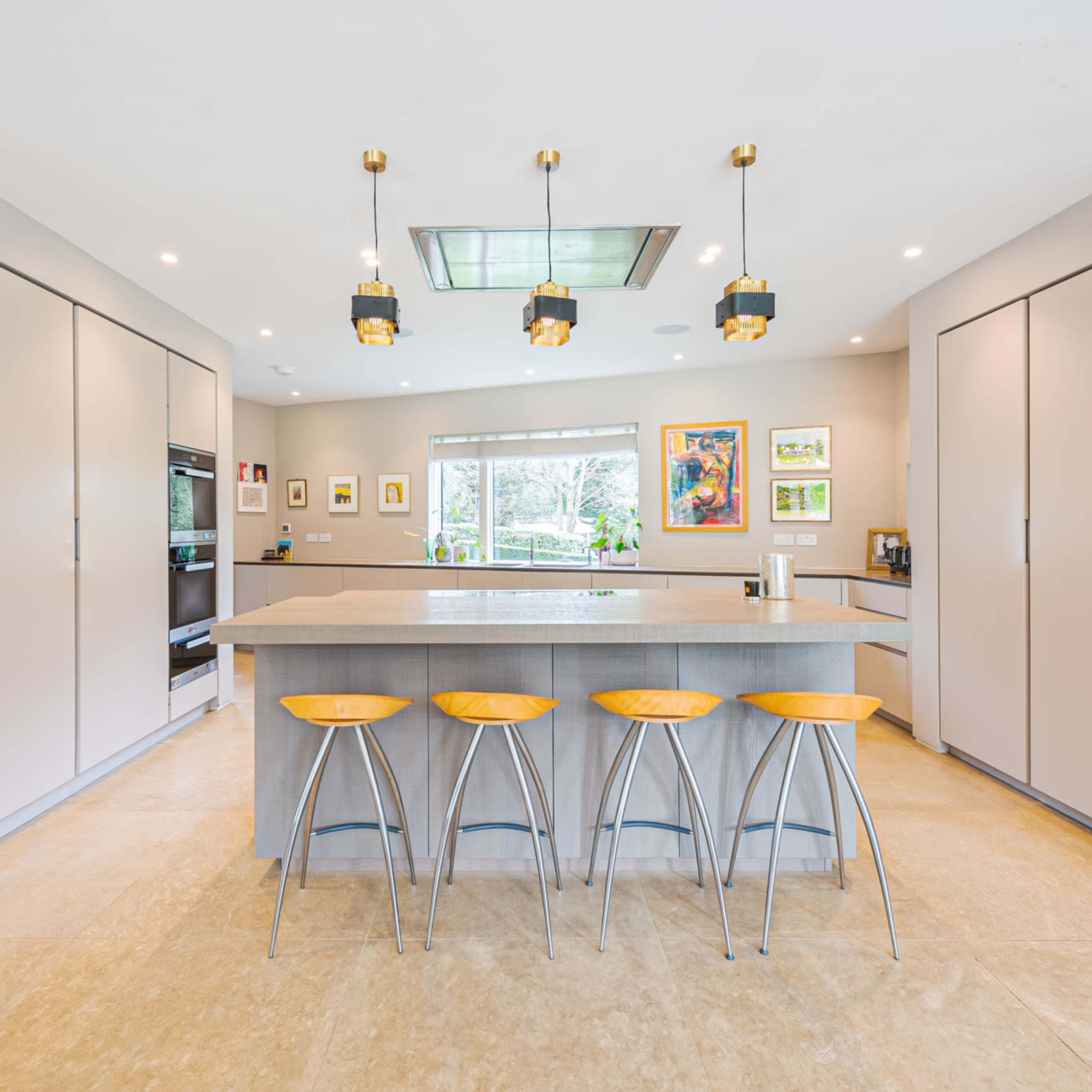 A modern kitchen with light gray cabinetry, a central island with four wooden bar stools, and large windows offering natural light.