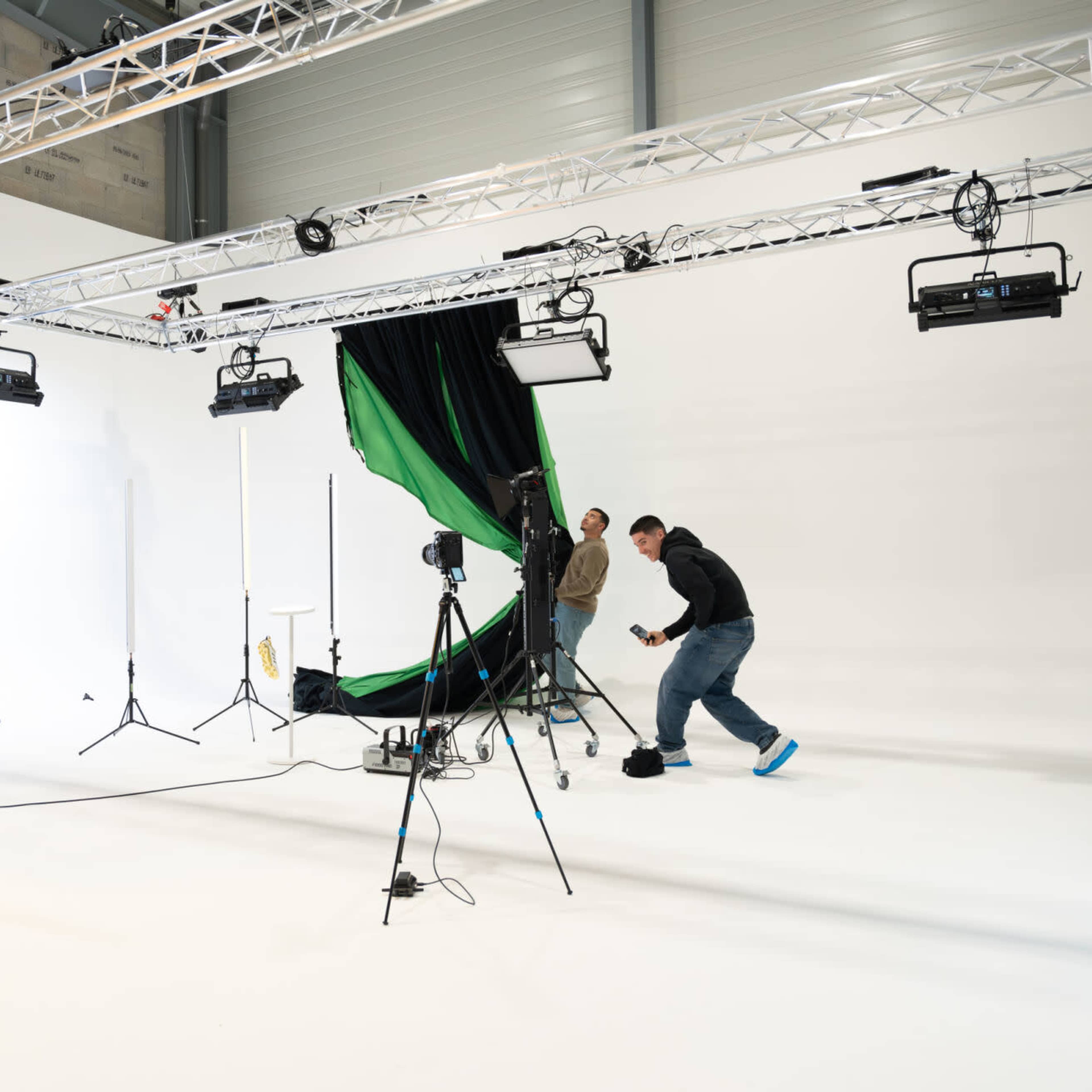 A photography studio with a white background is set up with lighting equipment, a green backdrop, and two individuals adjusting the scene.