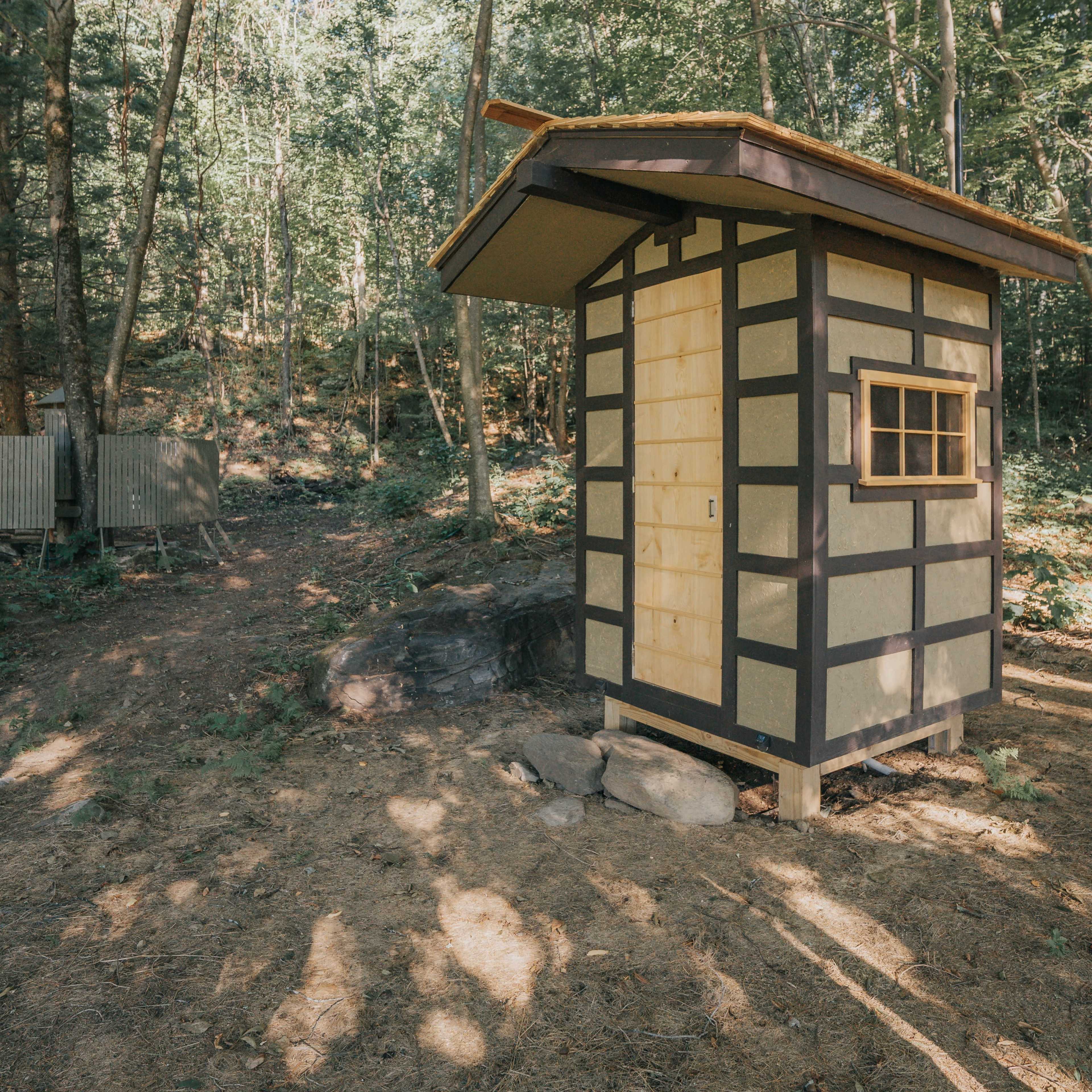 A small, wooden cabin with a slanted roof and a single window is surrounded by trees in a forested area.