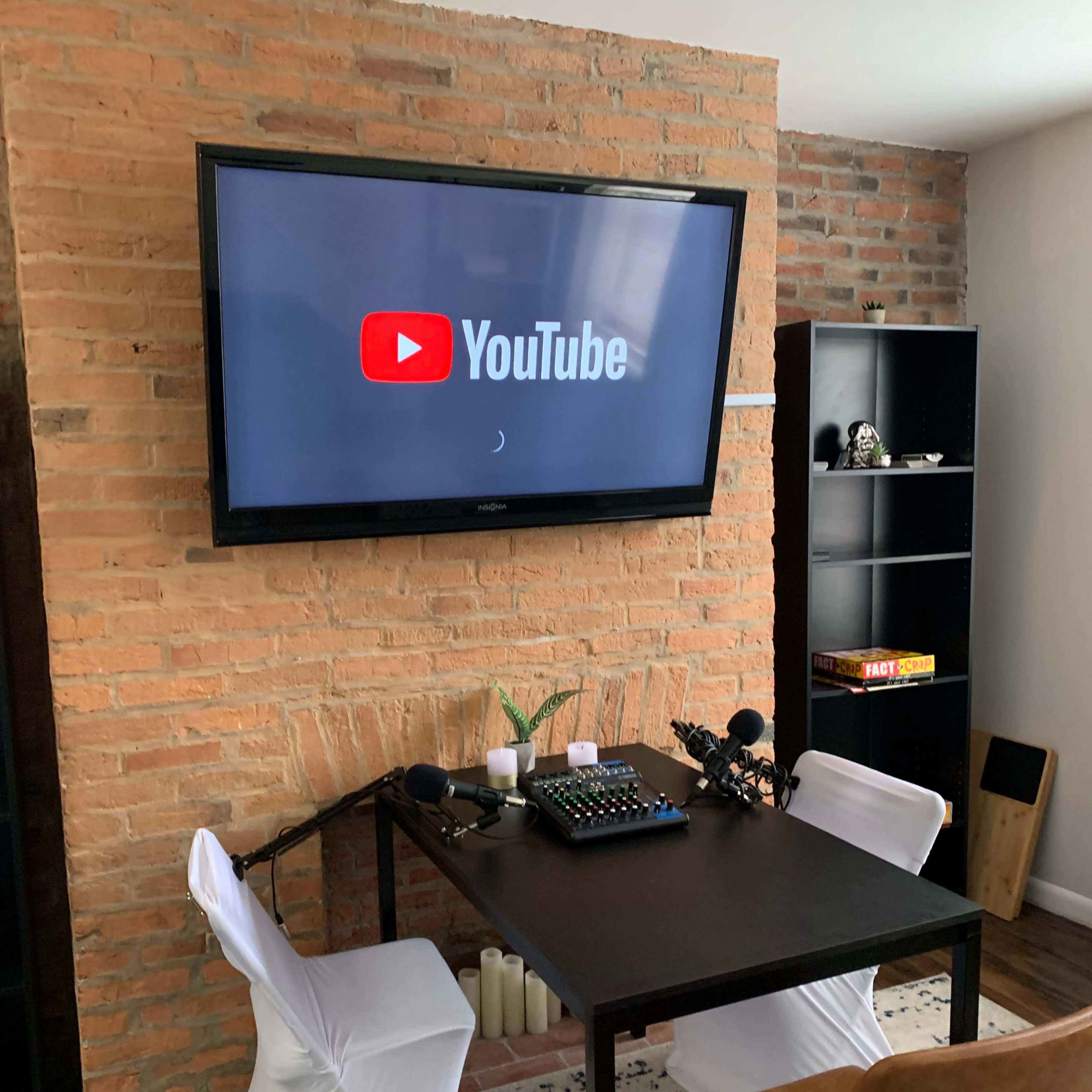 A black table with microphones and a control panel is positioned in front of a television displaying the YouTube logo against a brick wall.