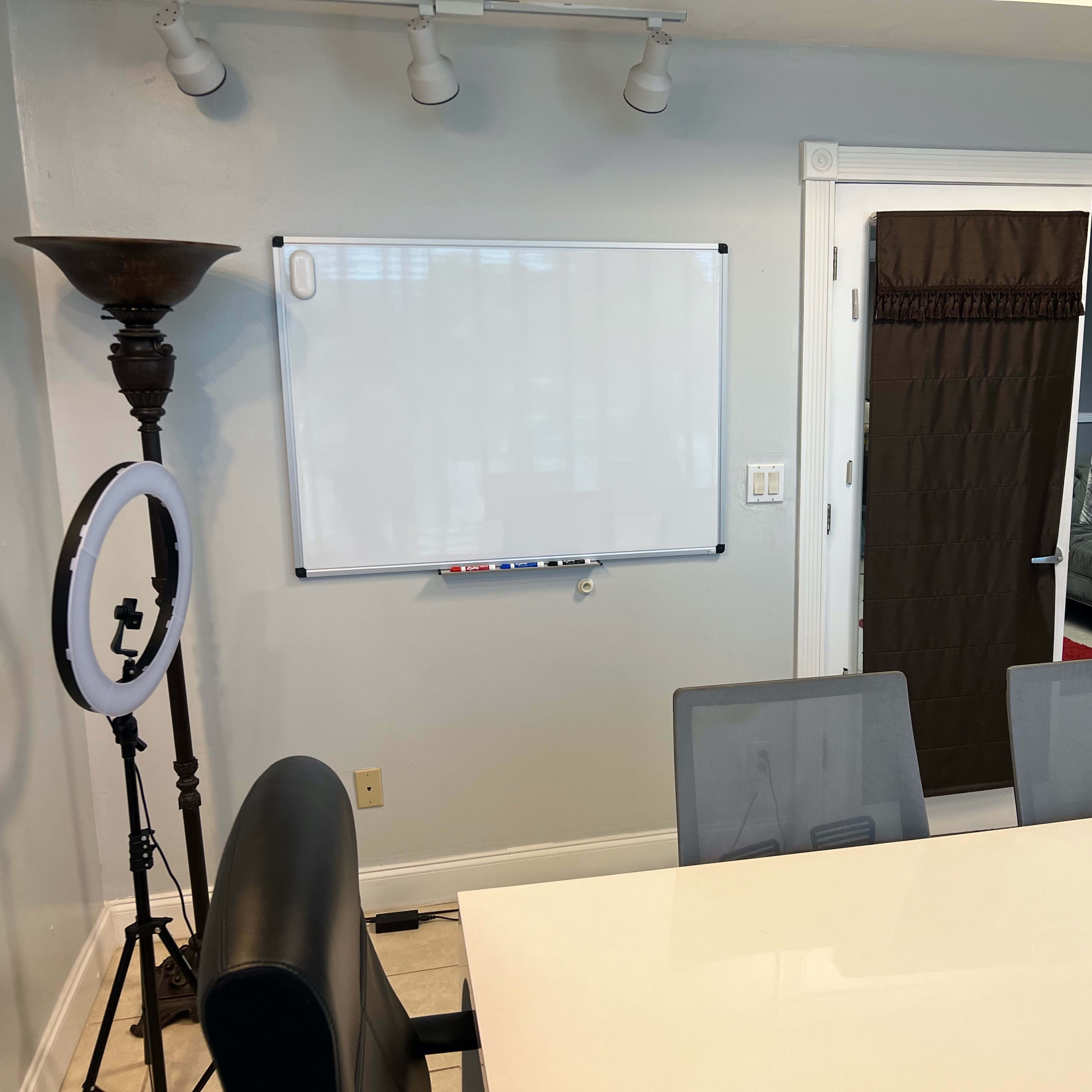 A meeting room with a whiteboard on the wall, a large table with chairs, and a ring light placed beside a floor lamp.