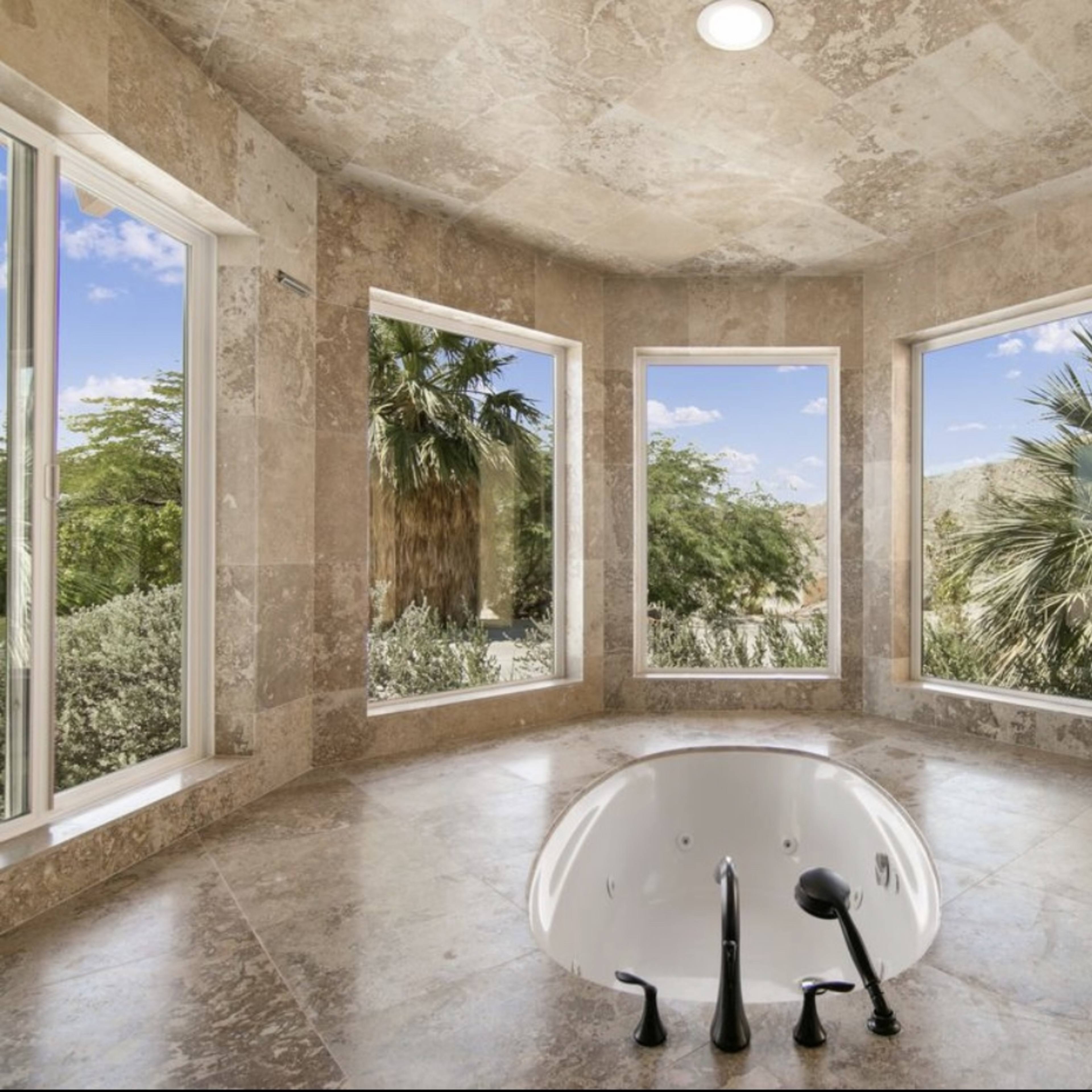 A soaking tub is positioned in a corner of a spacious bathroom featuring large windows that provide a view of surrounding palm trees and greenery.