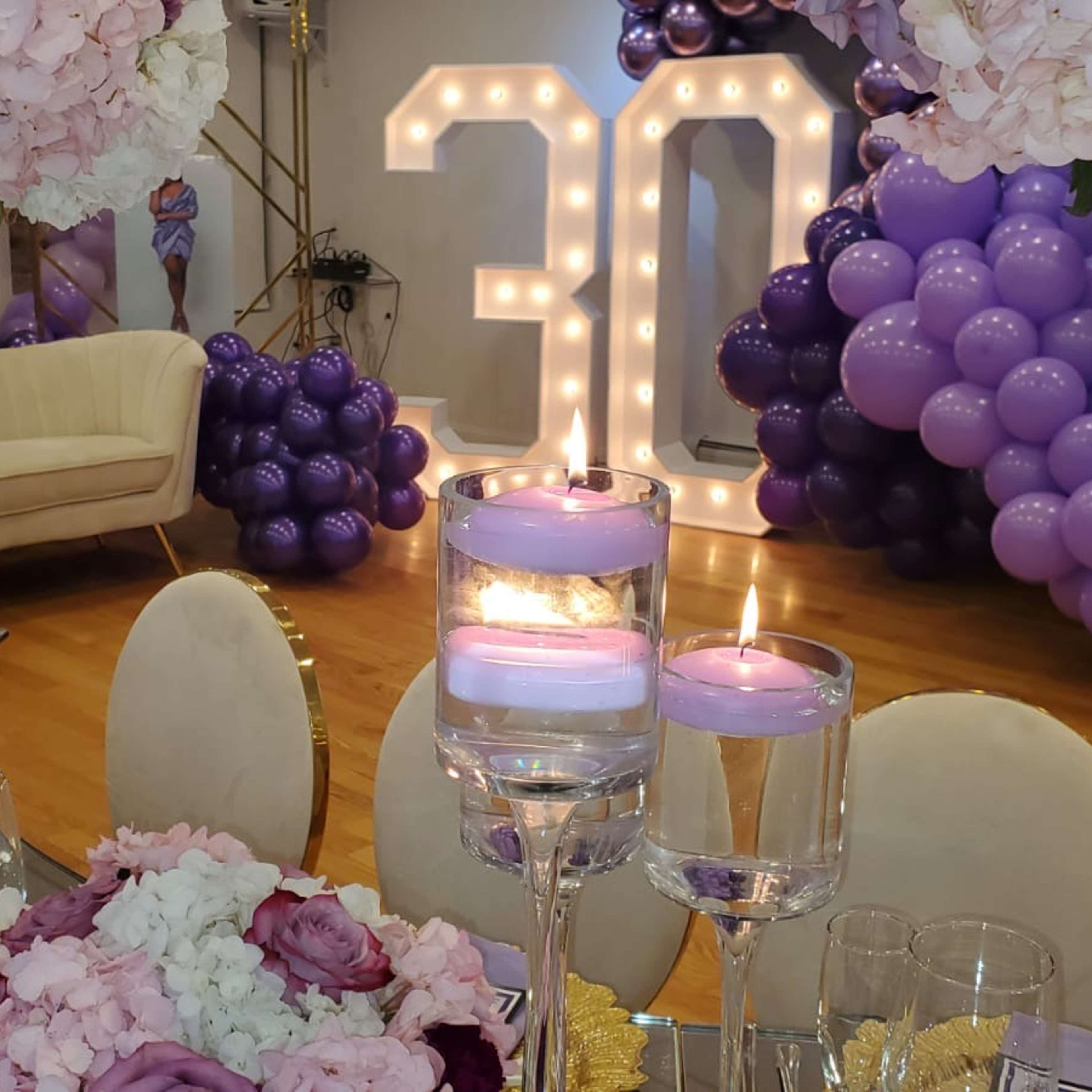 A elegantly decorated party space with large "30" illuminated numbers, a balloon arrangement in shades of purple, and floral centerpieces on dining tables.