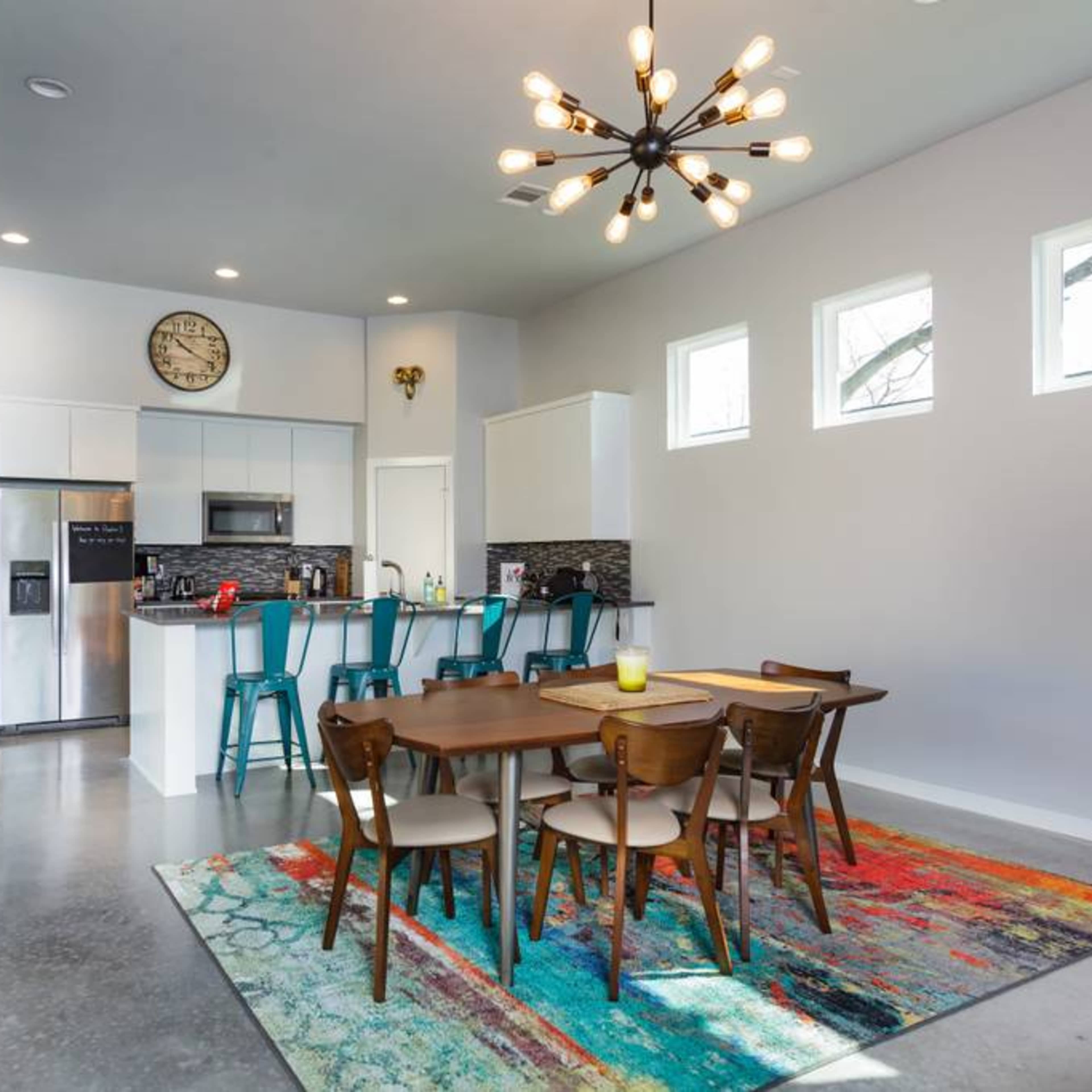 A modern kitchen and dining area with a large table, colorful rug, and contemporary lighting fixtures.