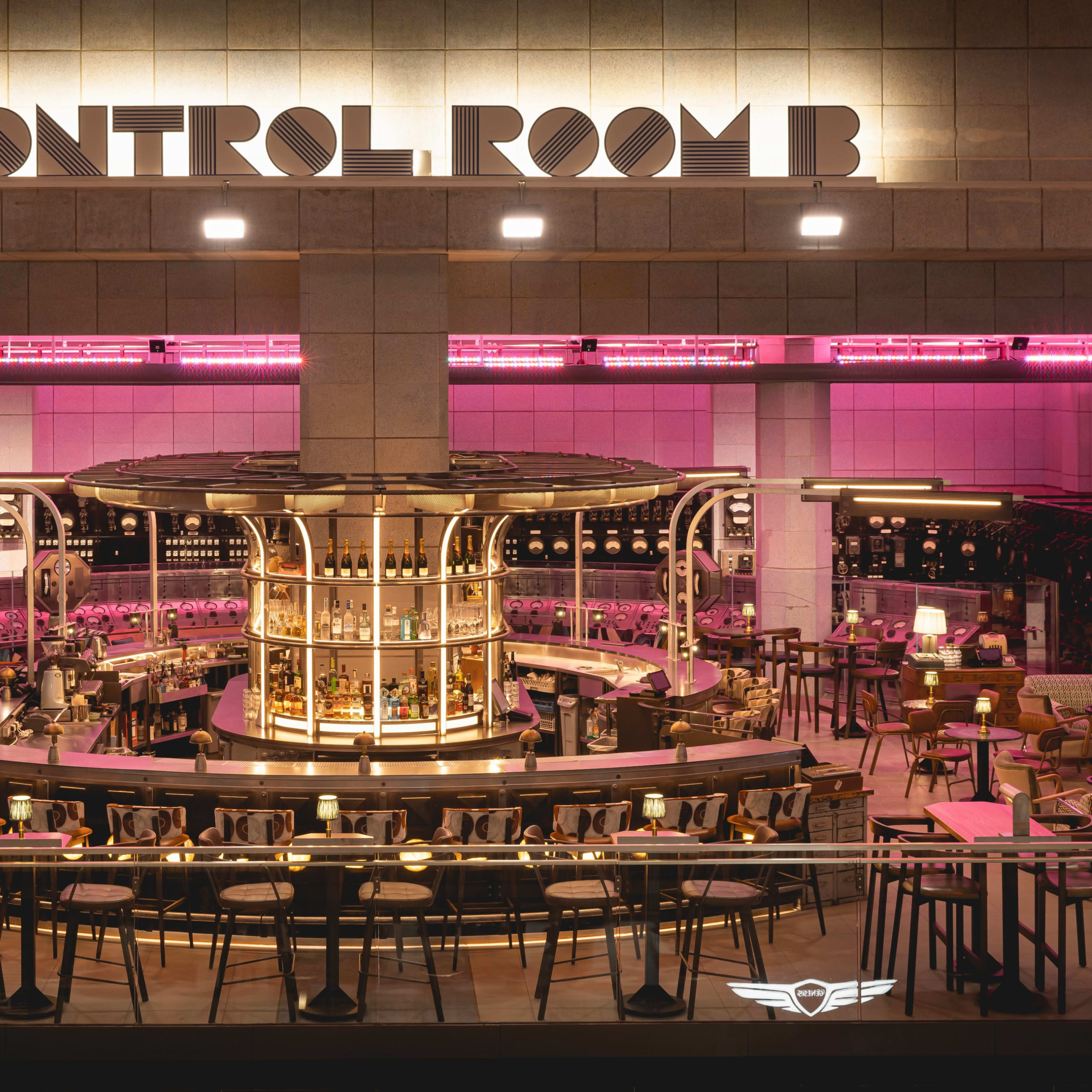 A bar area named "CONTROL ROOM B," featuring a circular bar surrounded by high stools, with pink lighting illuminating the space.