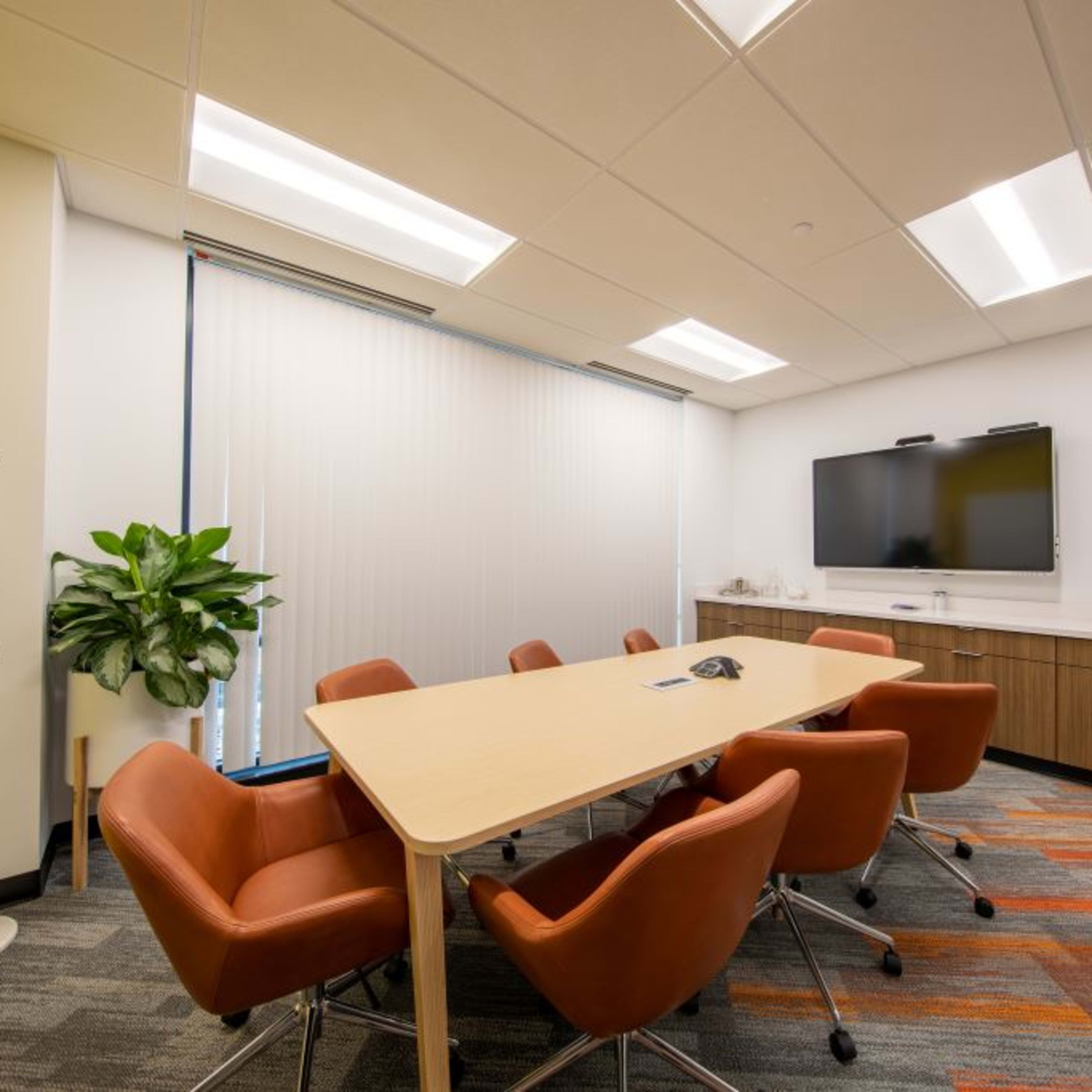 A conference room features a large table surrounded by orange chairs, a television mounted on the wall, and a potted plant in the corner.