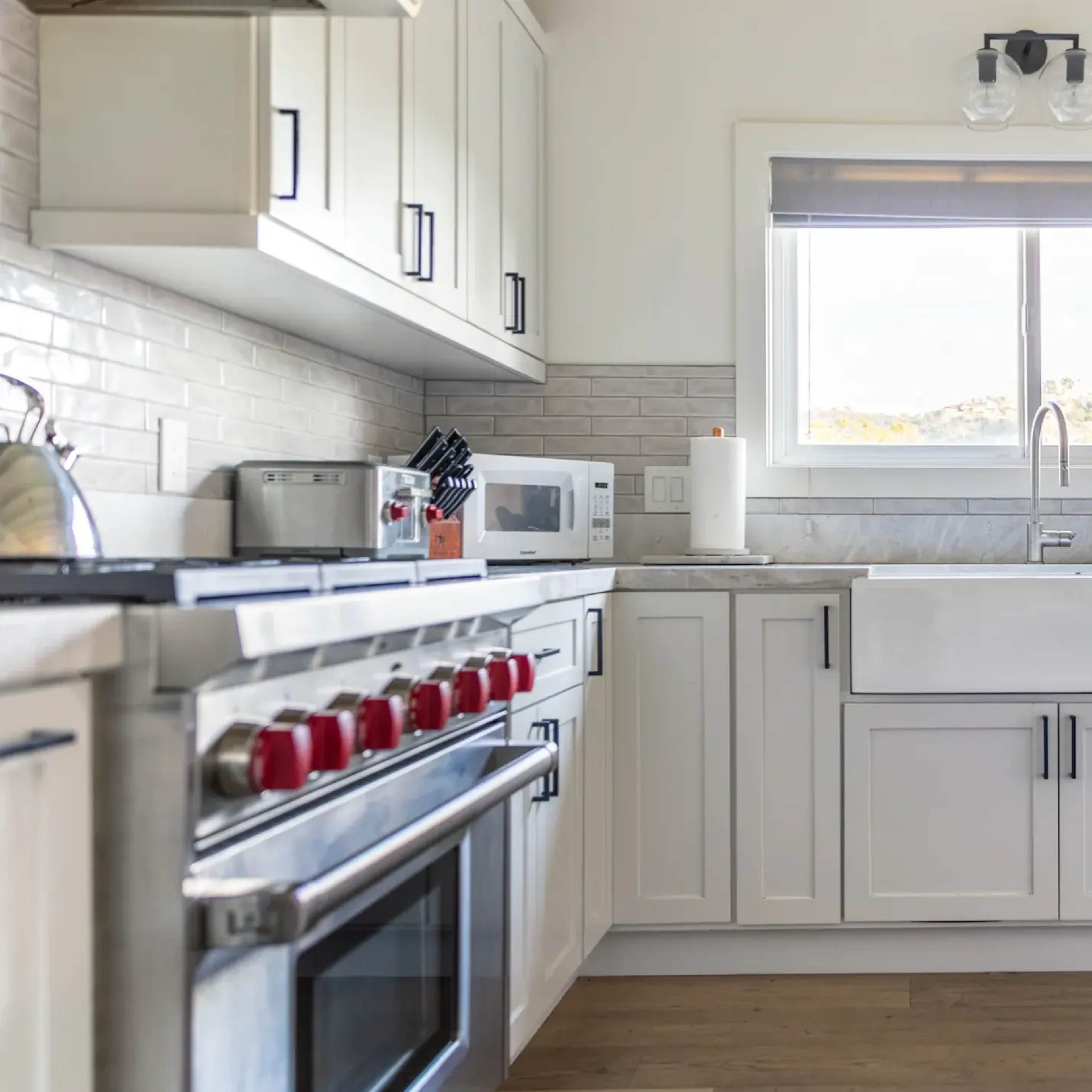 A modern kitchen features white cabinetry, a farmhouse sink, and stainless steel appliances, with a large window allowing natural light.