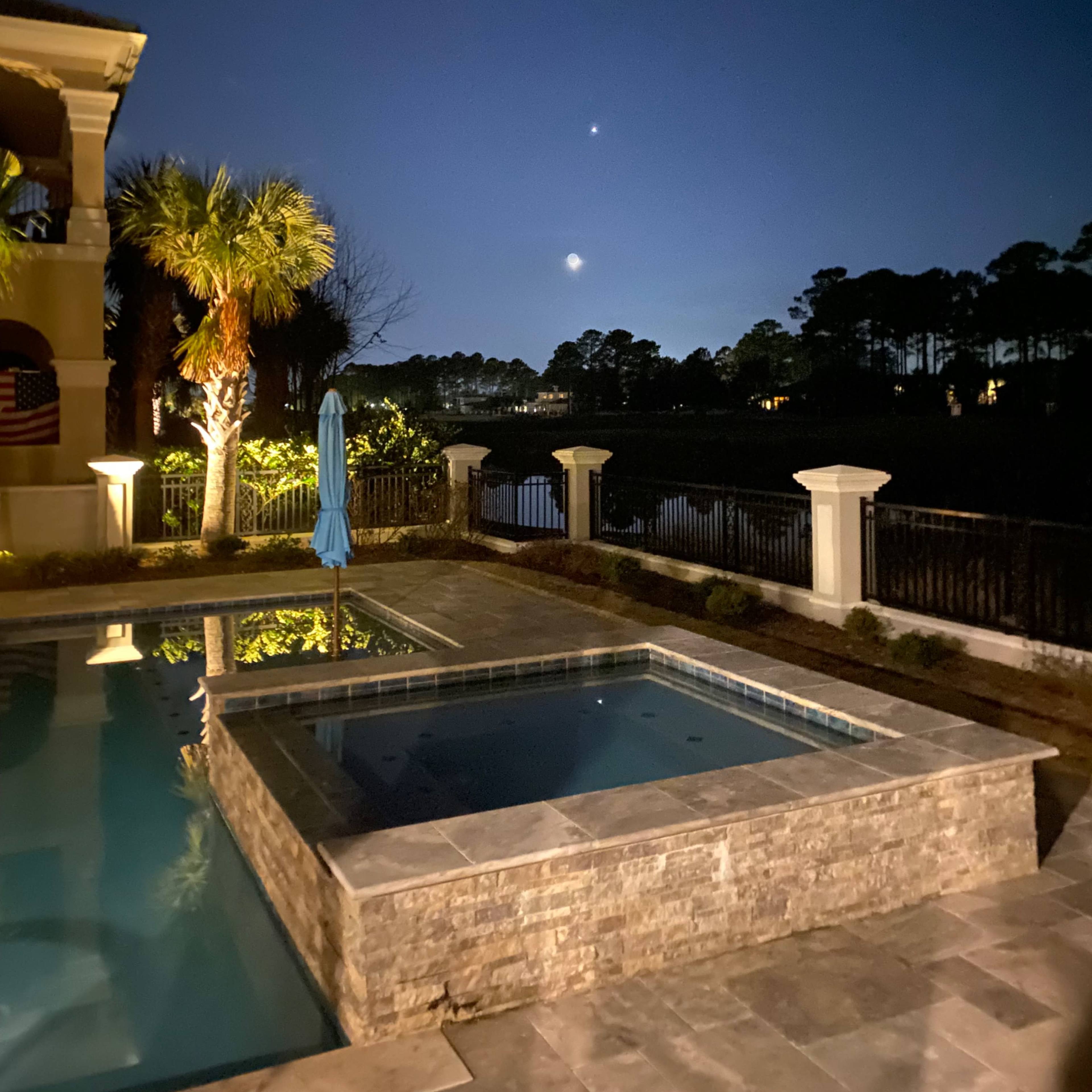 A backyard pool area at night, with a spa, a palm tree, and a view of the moon and two bright celestial objects in the sky.