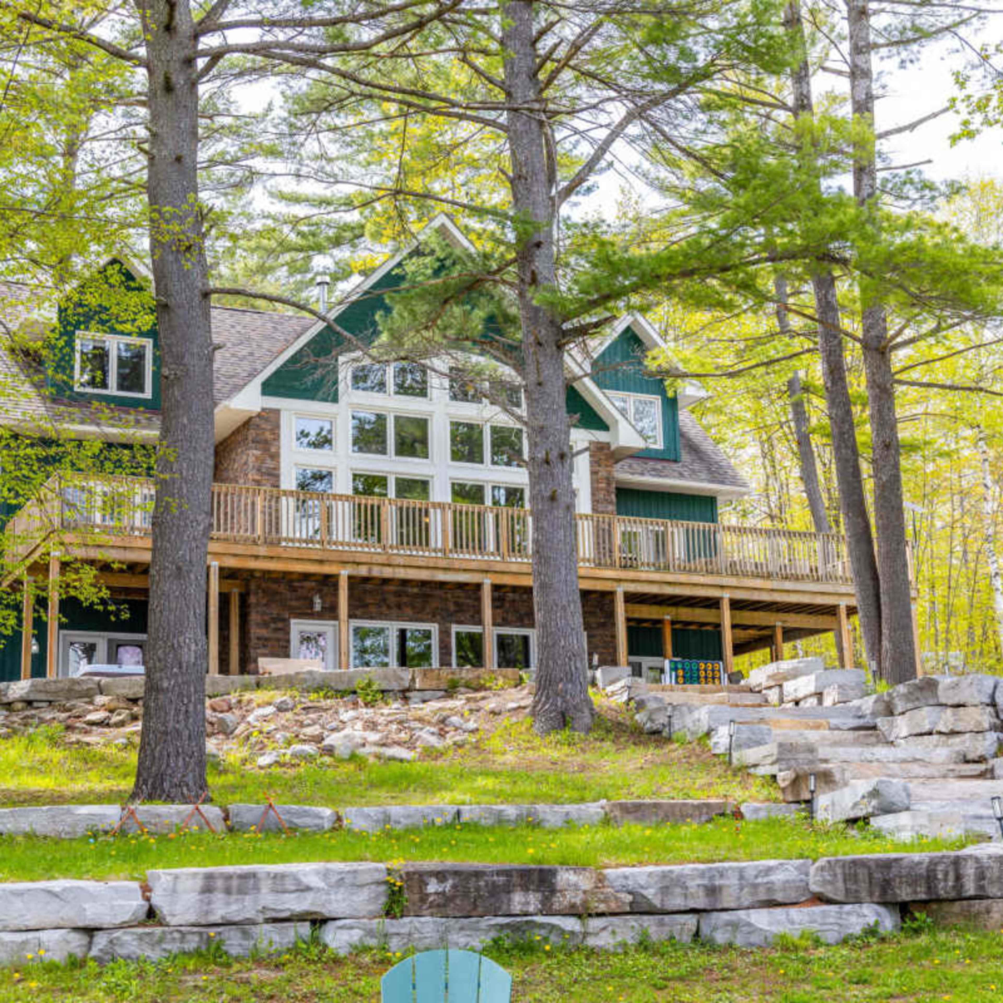 A large, multi-story house with a stone facade and expansive deck is nestled among tall trees and landscaped stone steps.