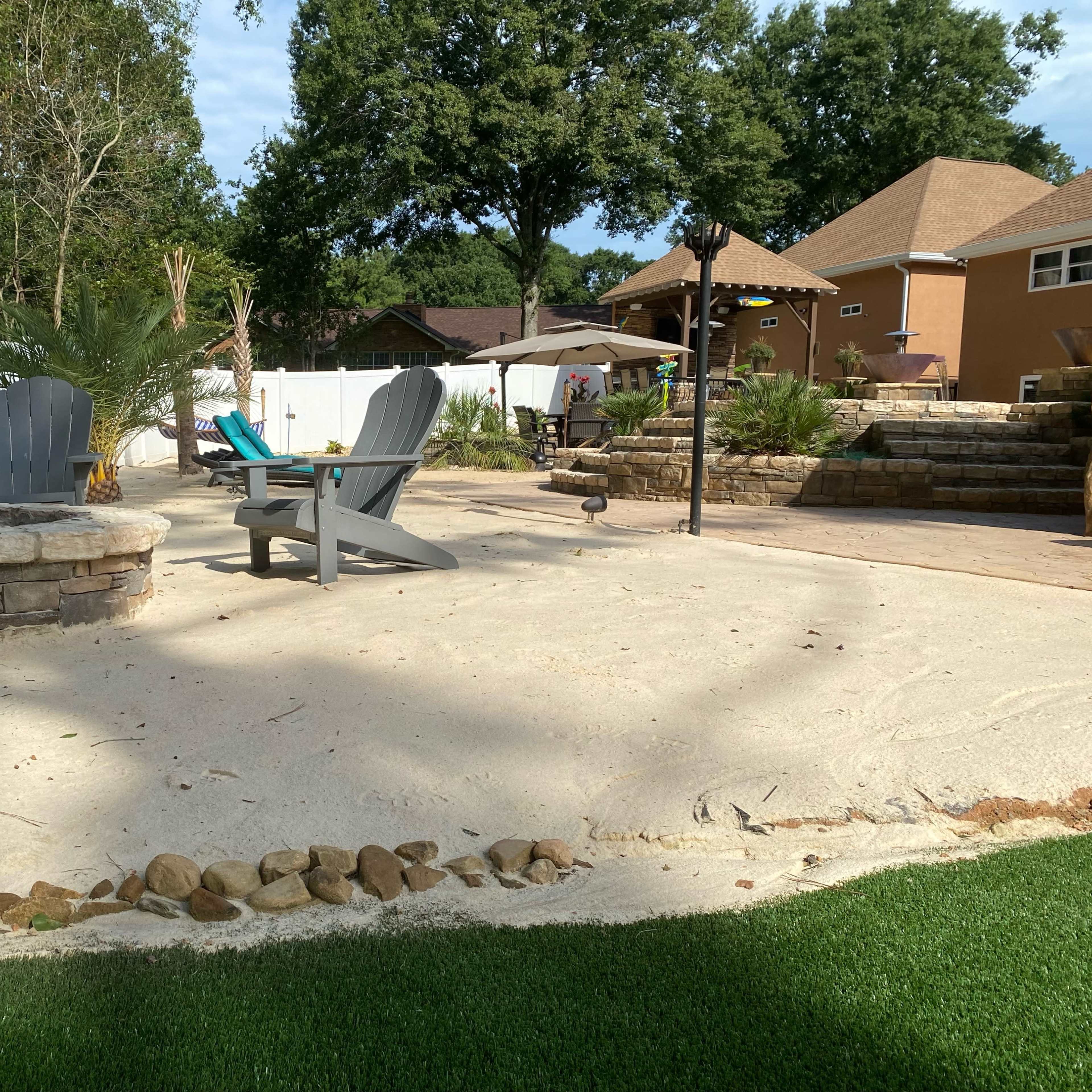 A landscaped backyard featuring a stone fire pit surrounded by two Adirondack chairs, with a sandy area and a green turf border.