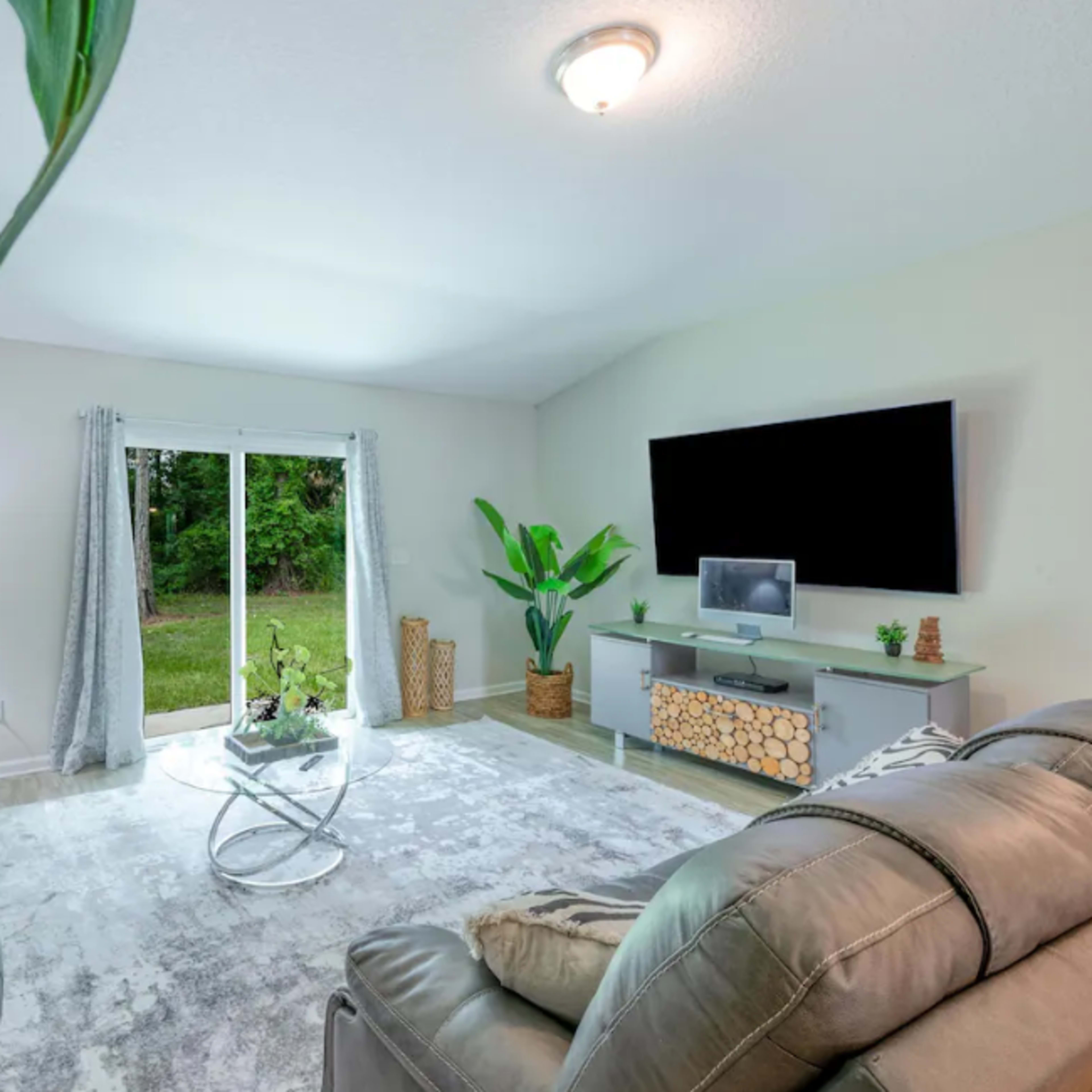 The living room features a pair of grey sofas, a glass coffee table, and a large television mounted on a light-colored wall, with windows overlooking a green outdoor area.