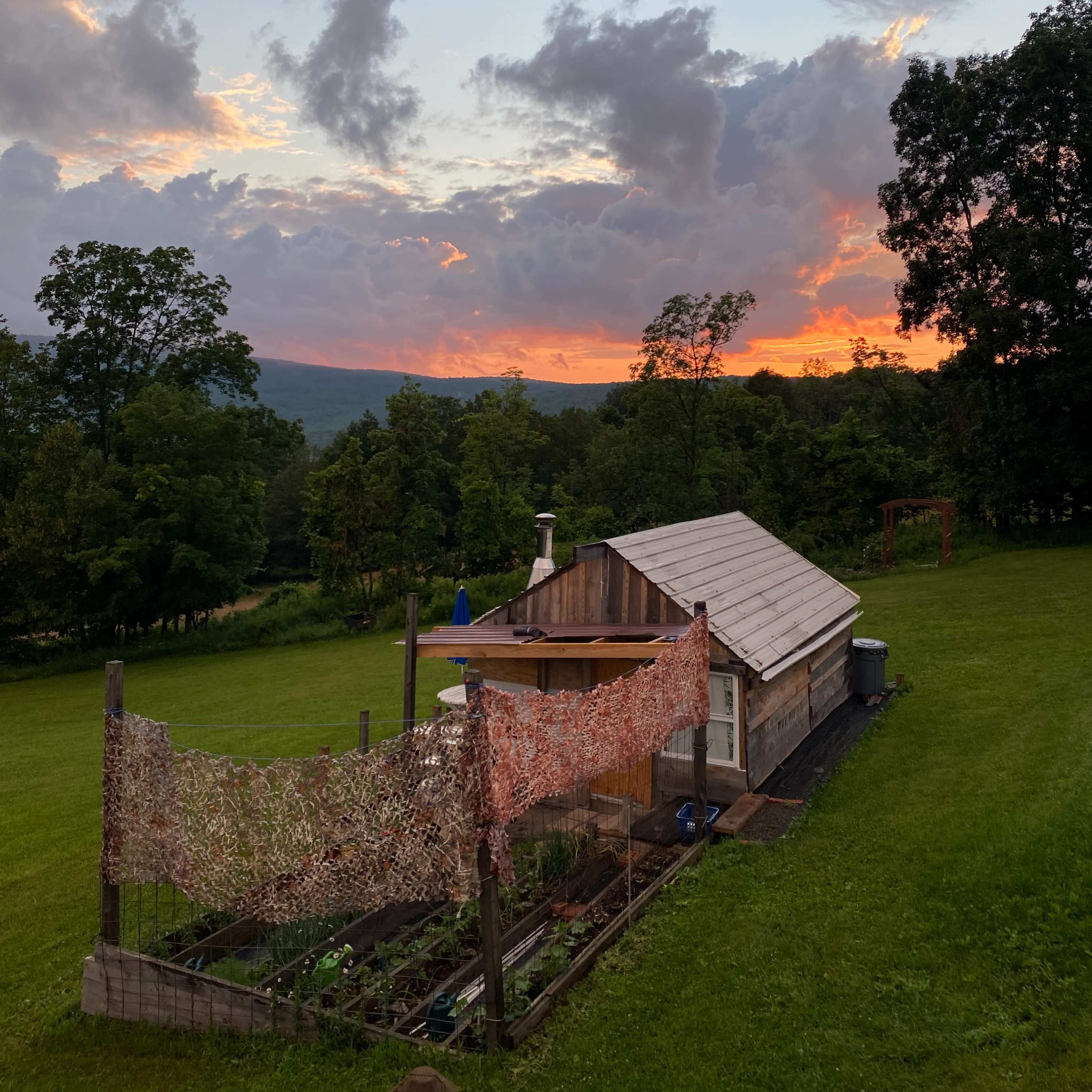 A small wooden cabin sits on green grass, surrounded by trees, with a colorful sunset and clouds overhead.