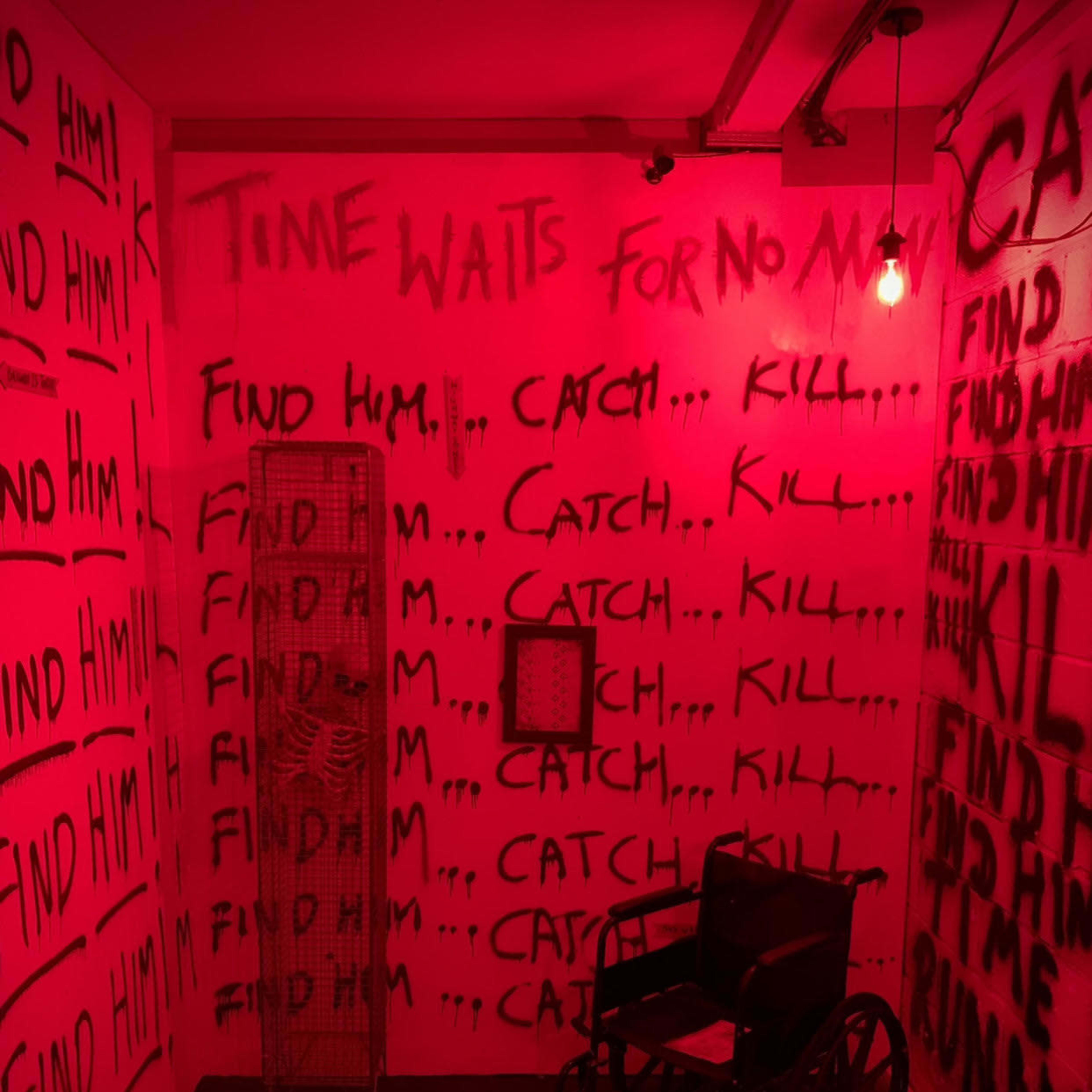 A dimly lit room with red walls covered in graffiti that reads phrases like "FIND HIM," "CATCH," and "KILL," alongside an empty wheelchair and a wire cage.