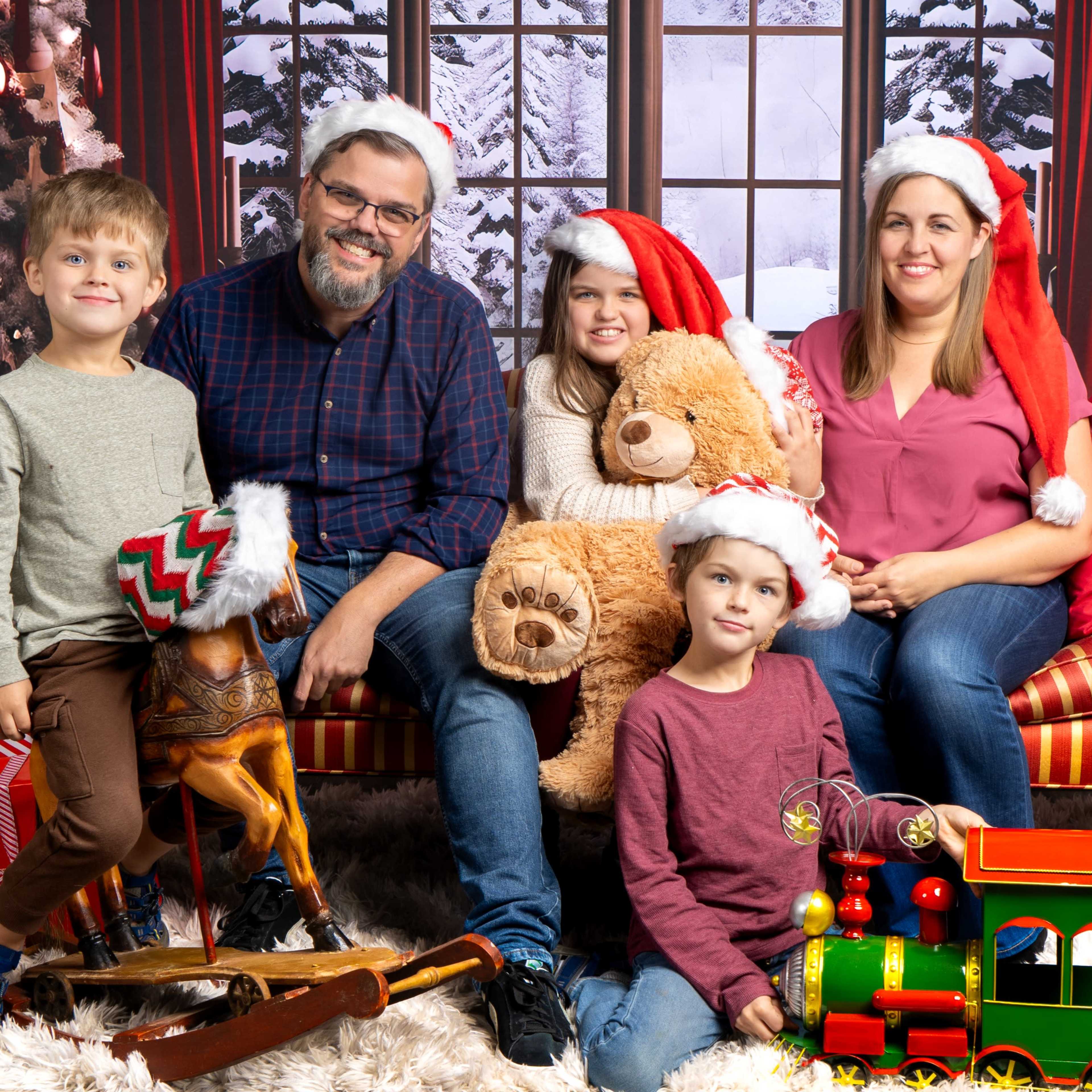 A family of five wearing festive hats poses on a couch surrounded by holiday decorations and gifts.