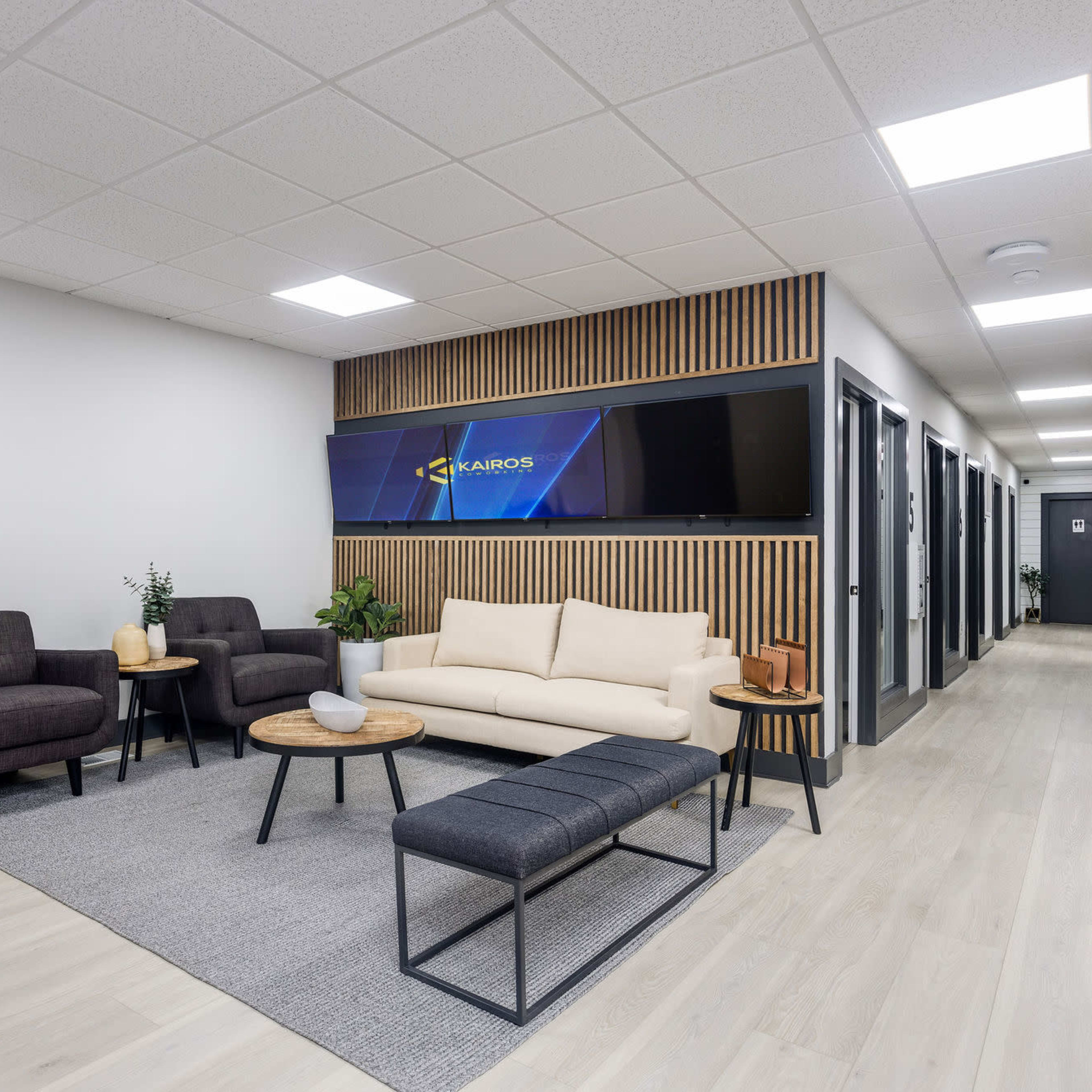 A modern office waiting area with grey sofas, a coffee table, and a large screen mounted on a wooden panel against the wall, leading to a hallway with multiple doors.