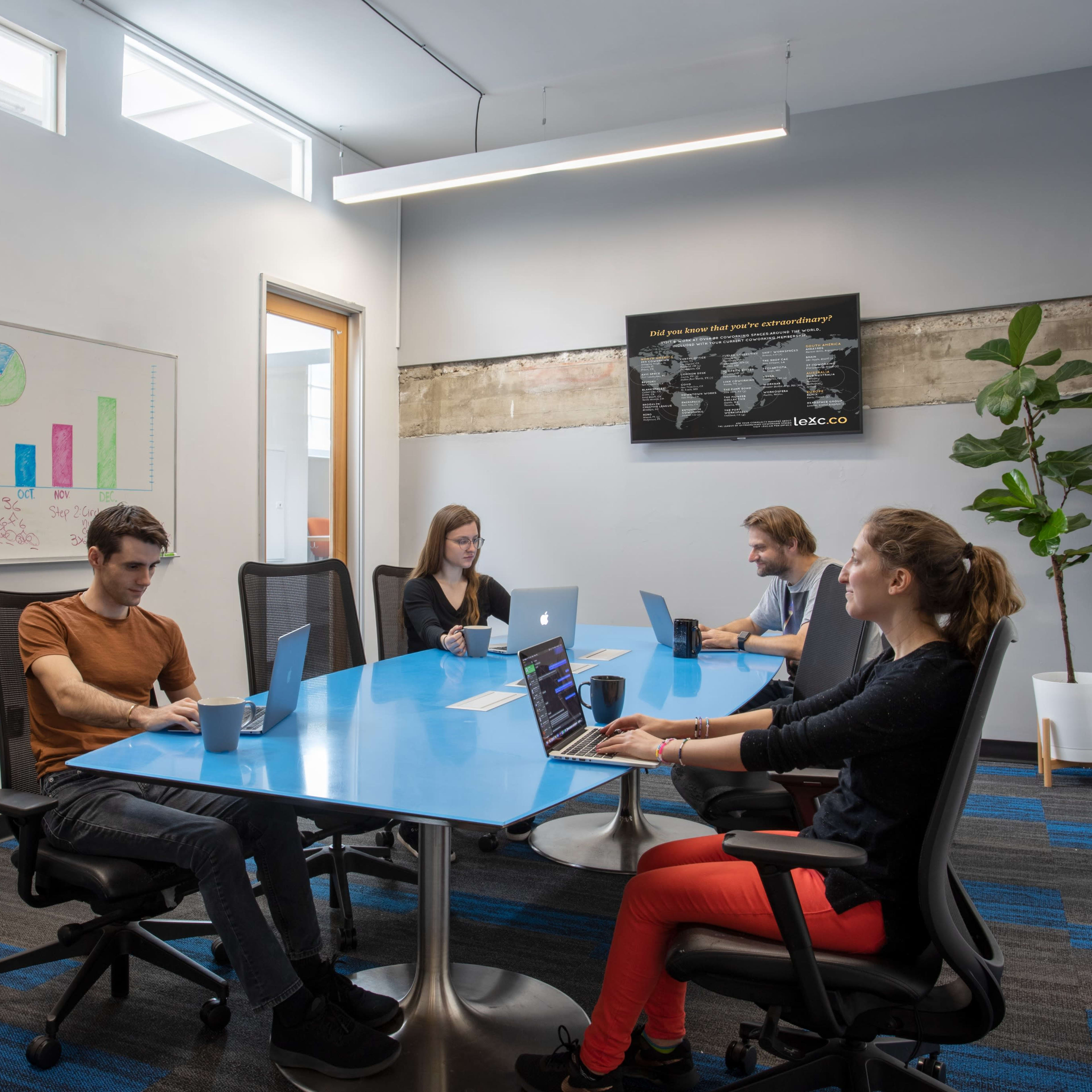 A group of four people work at a blue conference table in a modern office, with laptops and a whiteboard in the background.
