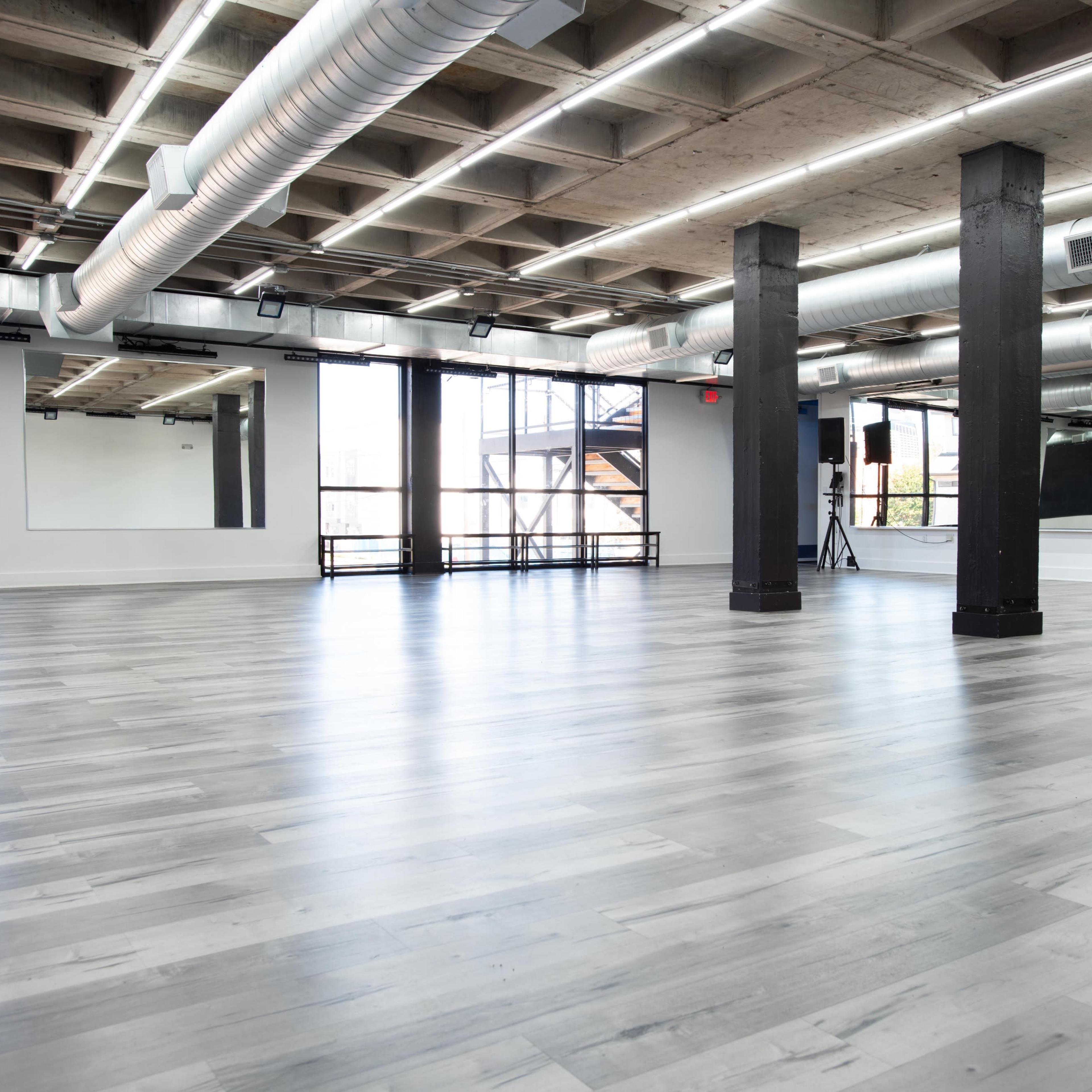 A spacious, modern studio with large windows, polished wooden floors, and exposed ductwork.
