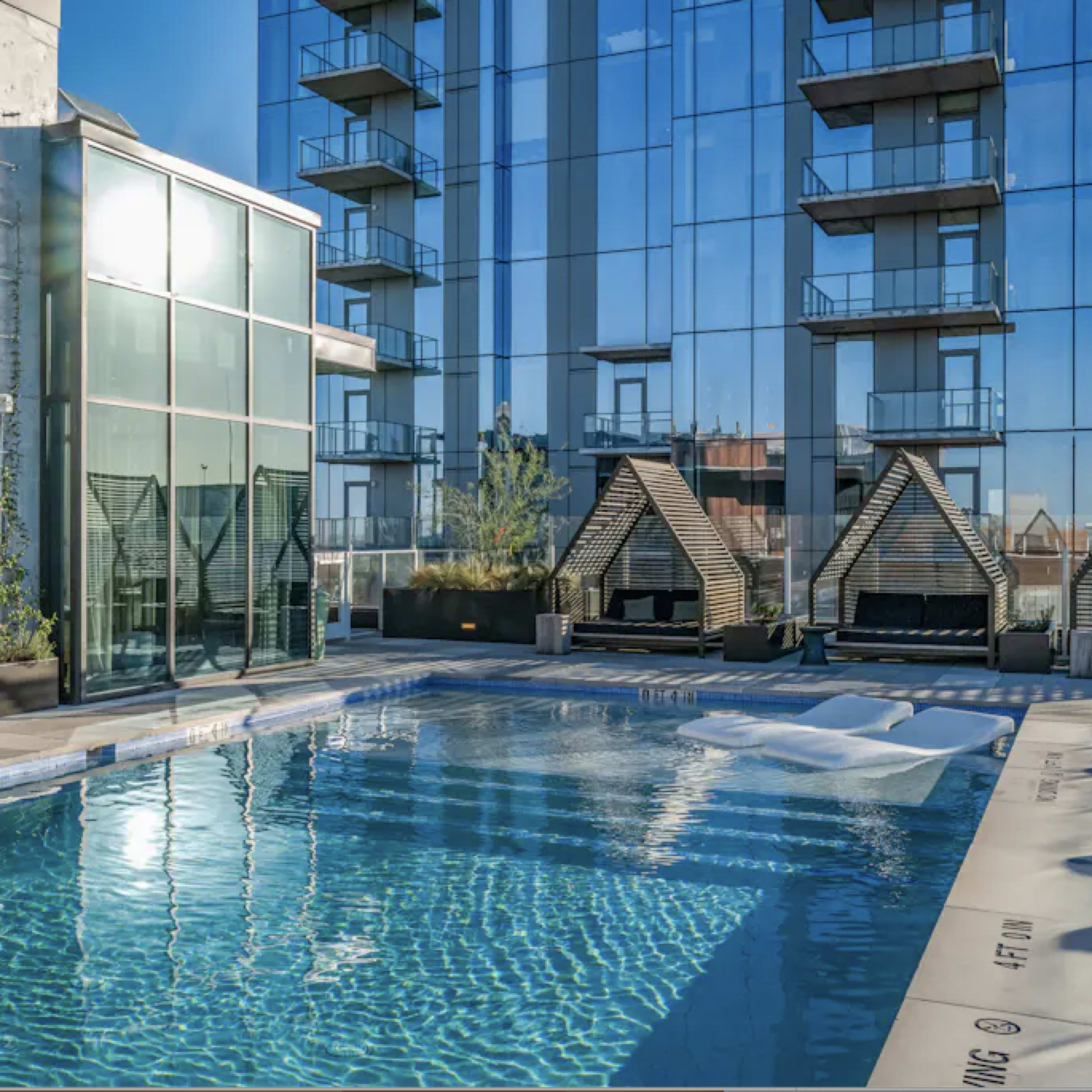 A modern swimming pool surrounded by lounge areas and a glass building reflecting in the water.