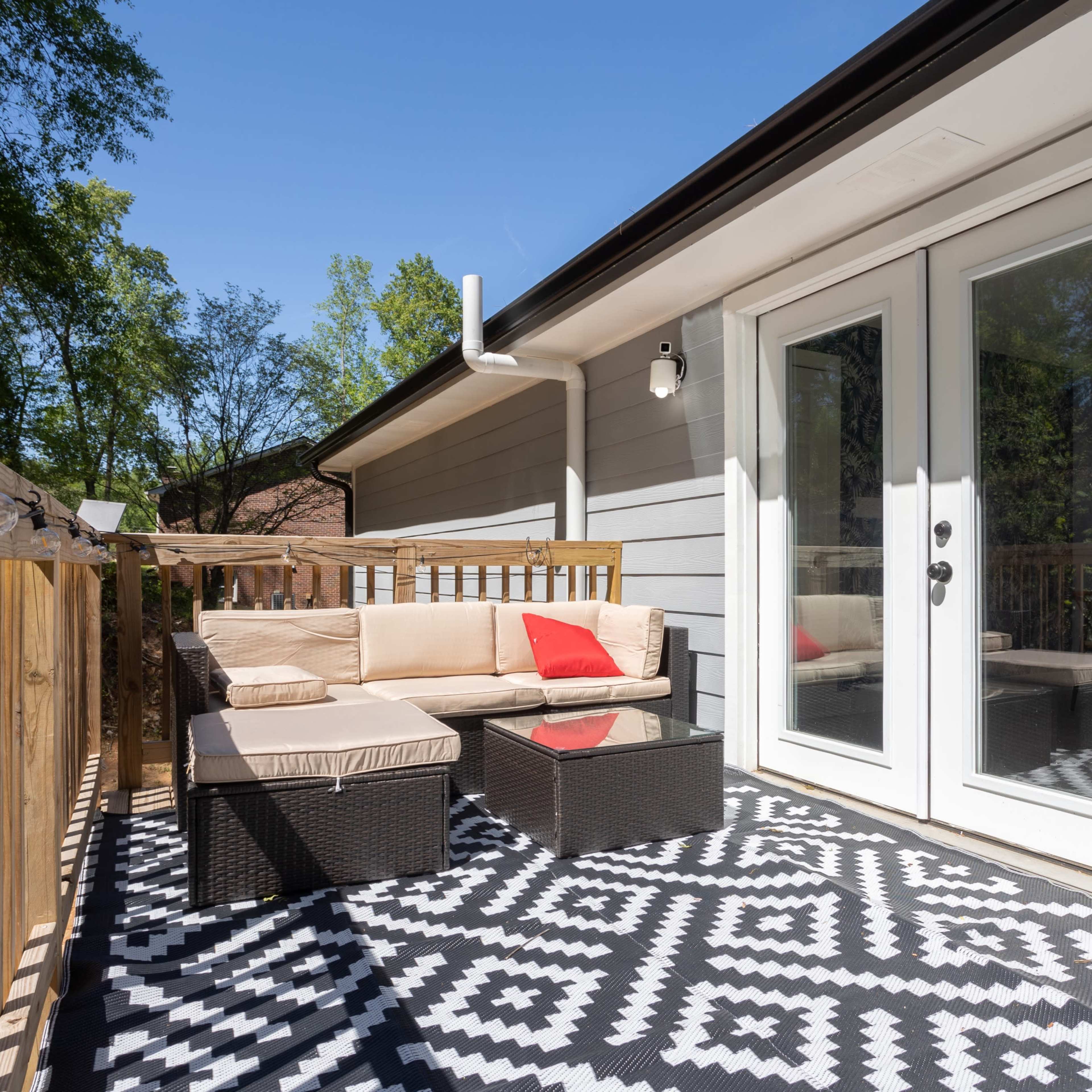 A deck features a seating area with a beige couch and a red cushion, accompanied by a coffee table, under clear blue skies.