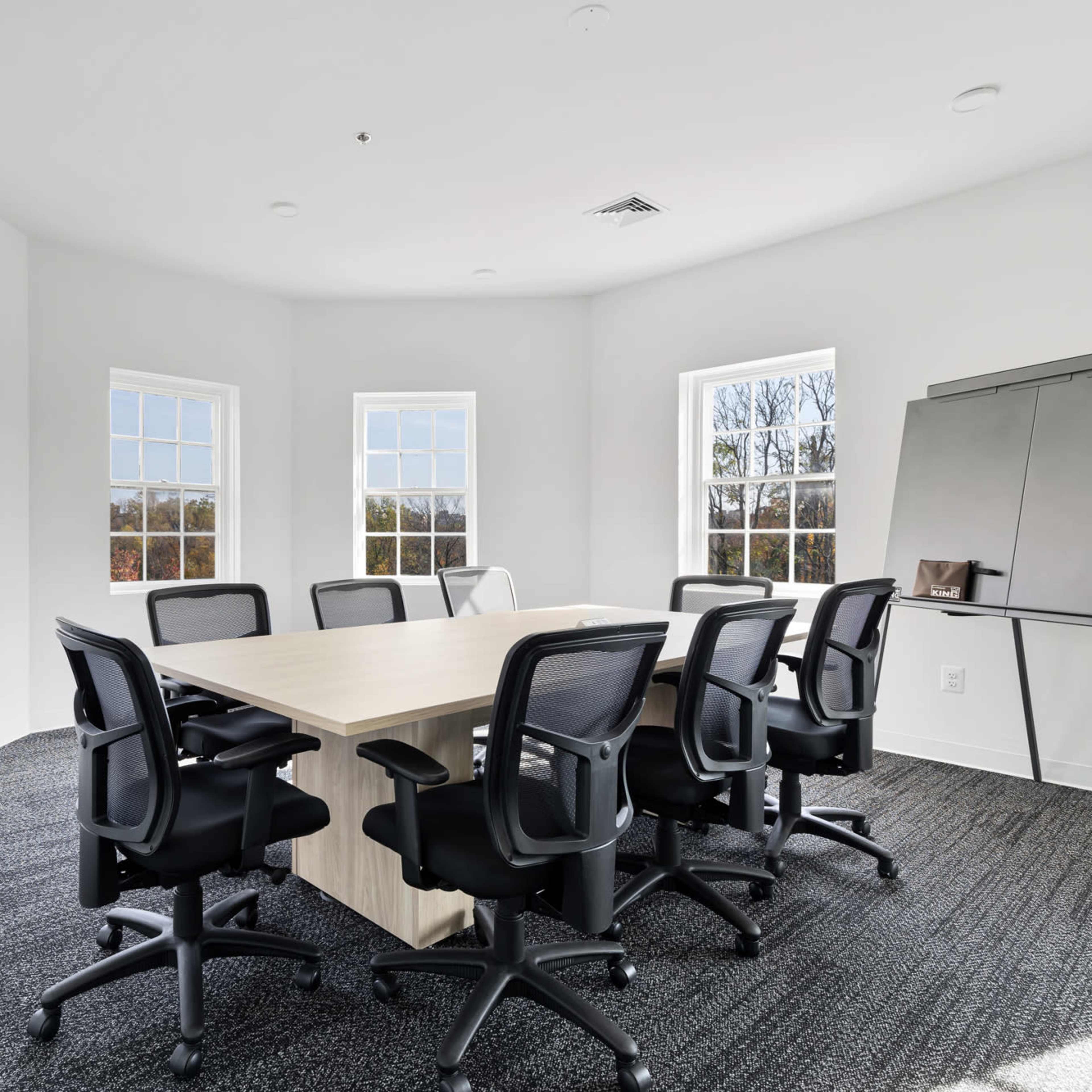 A conference room features a large table surrounded by black chairs, with windows on three walls allowing natural light to enter.