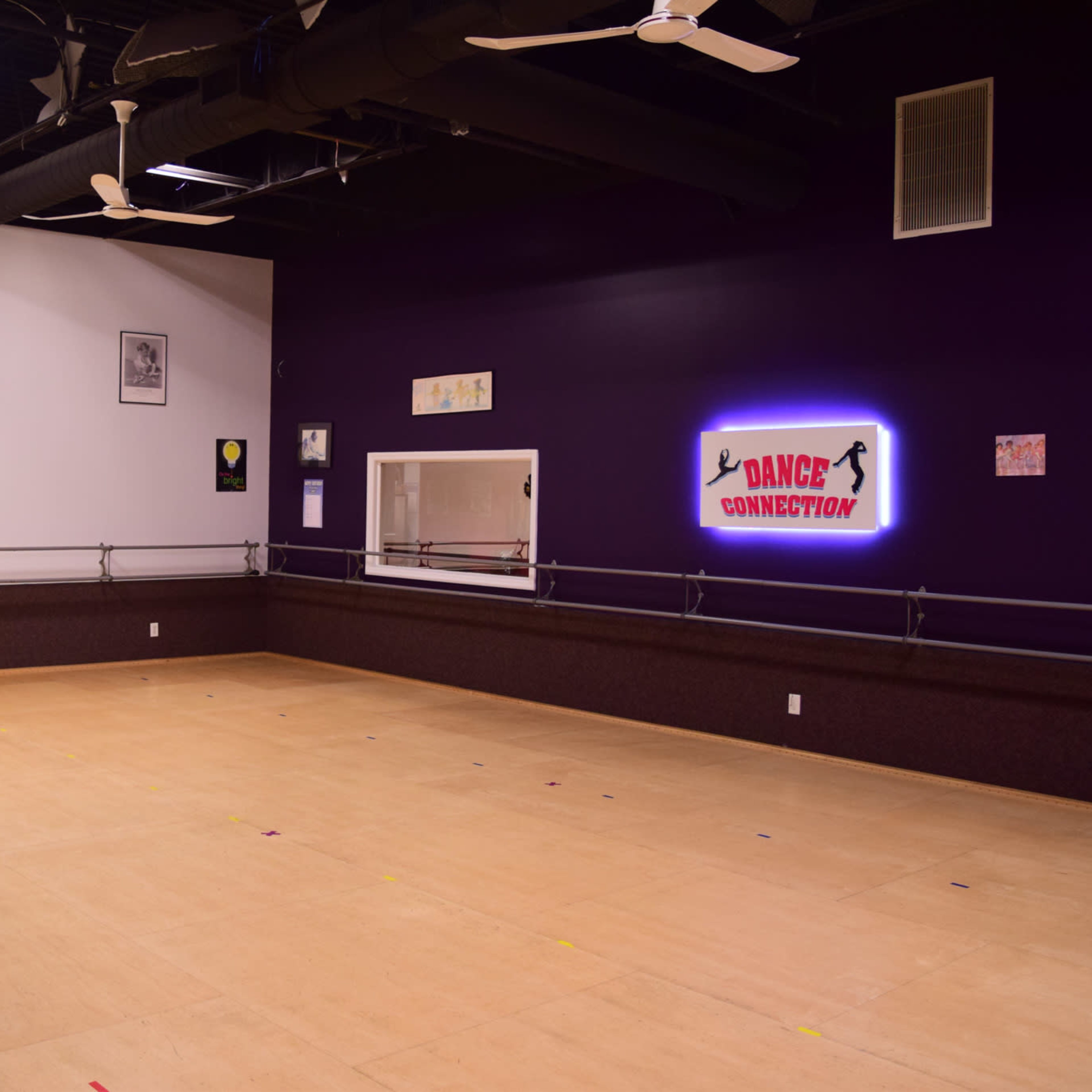 A spacious dance studio with a wooden floor, mirrored wall, and purple-painted walls, featuring the name "Dance Connection" illuminated on one side.