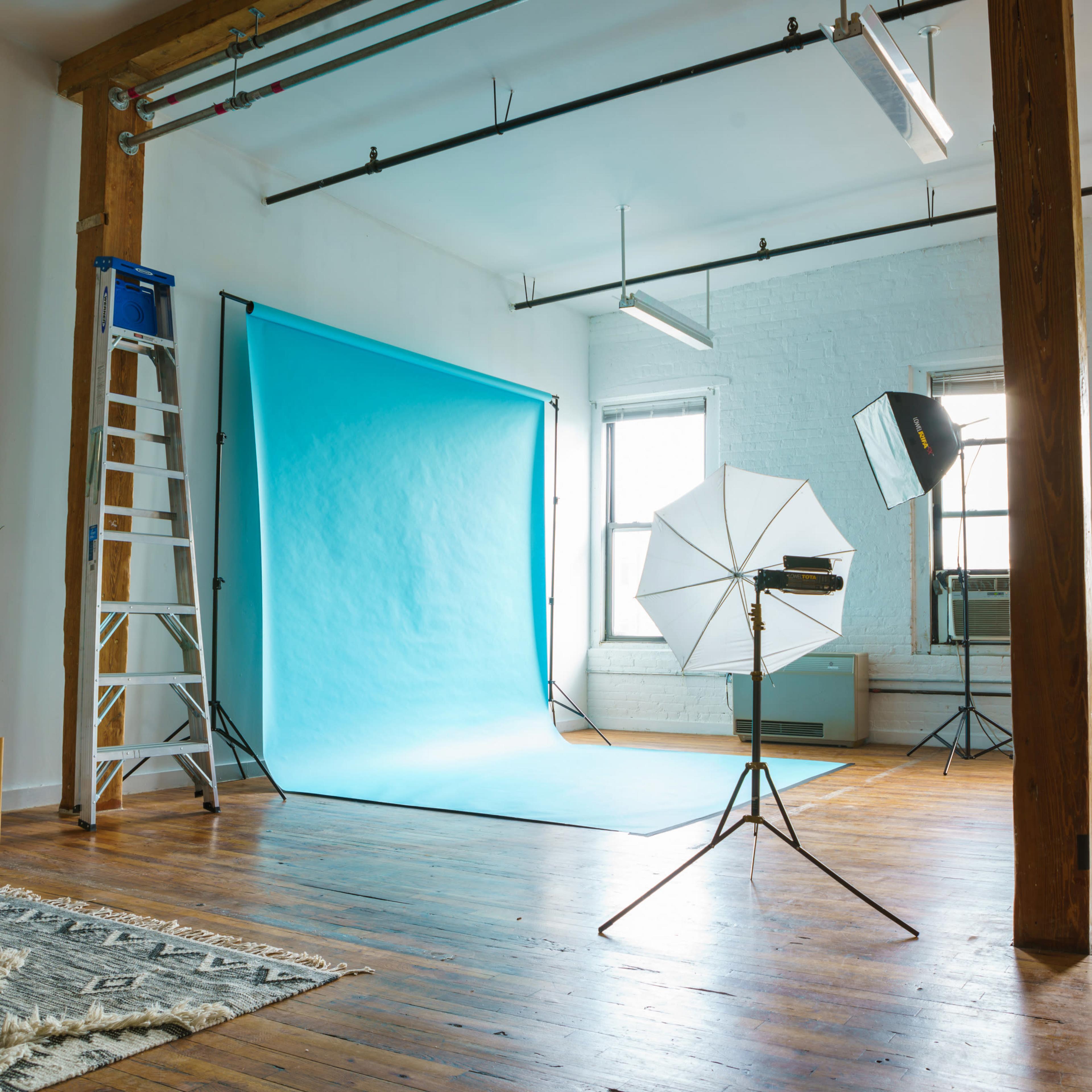 A photography studio with a blue backdrop, lighting equipment, a ladder, and wooden flooring.