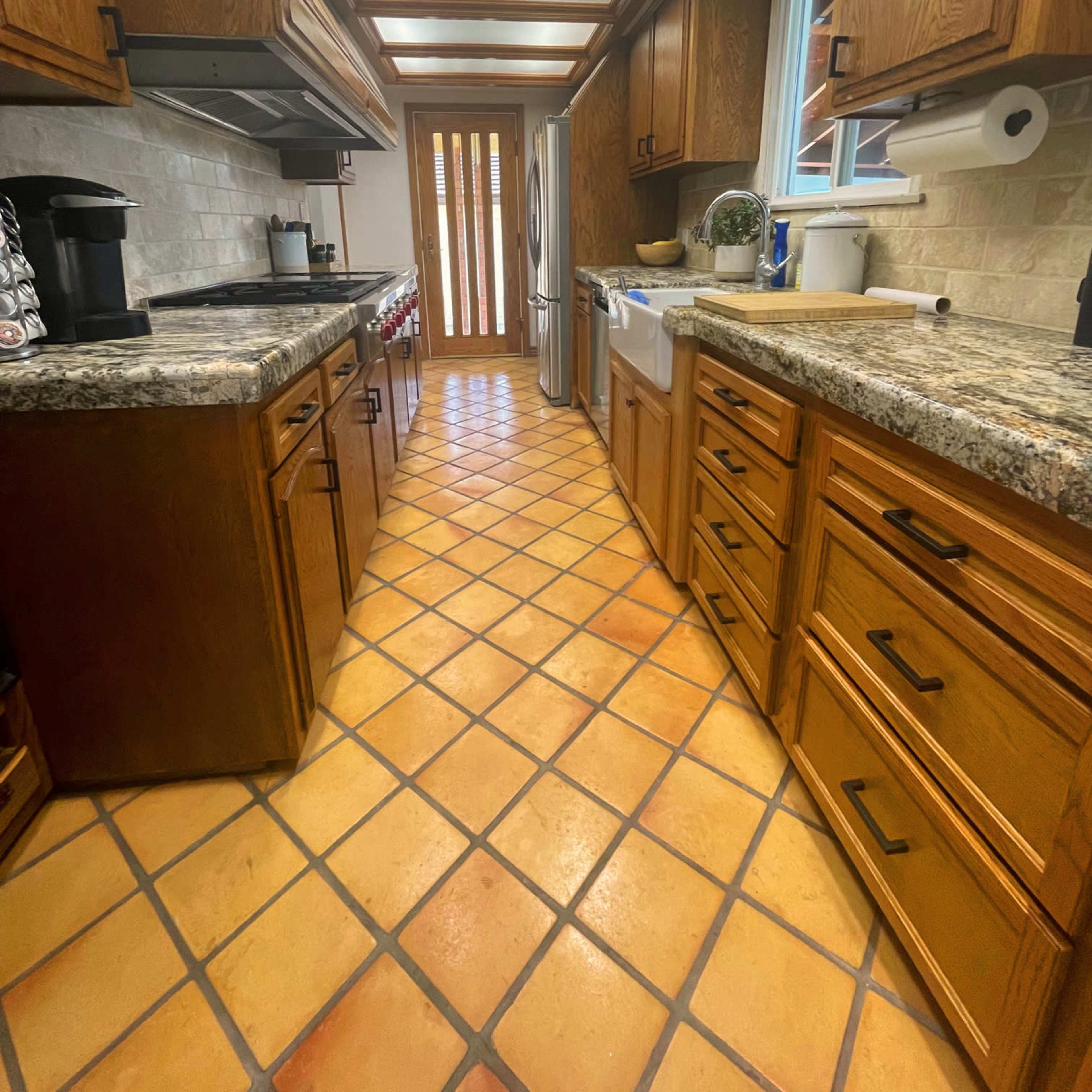 A narrow kitchen with wooden cabinets, granite countertops, and a tiled floor leading to a door at the end.