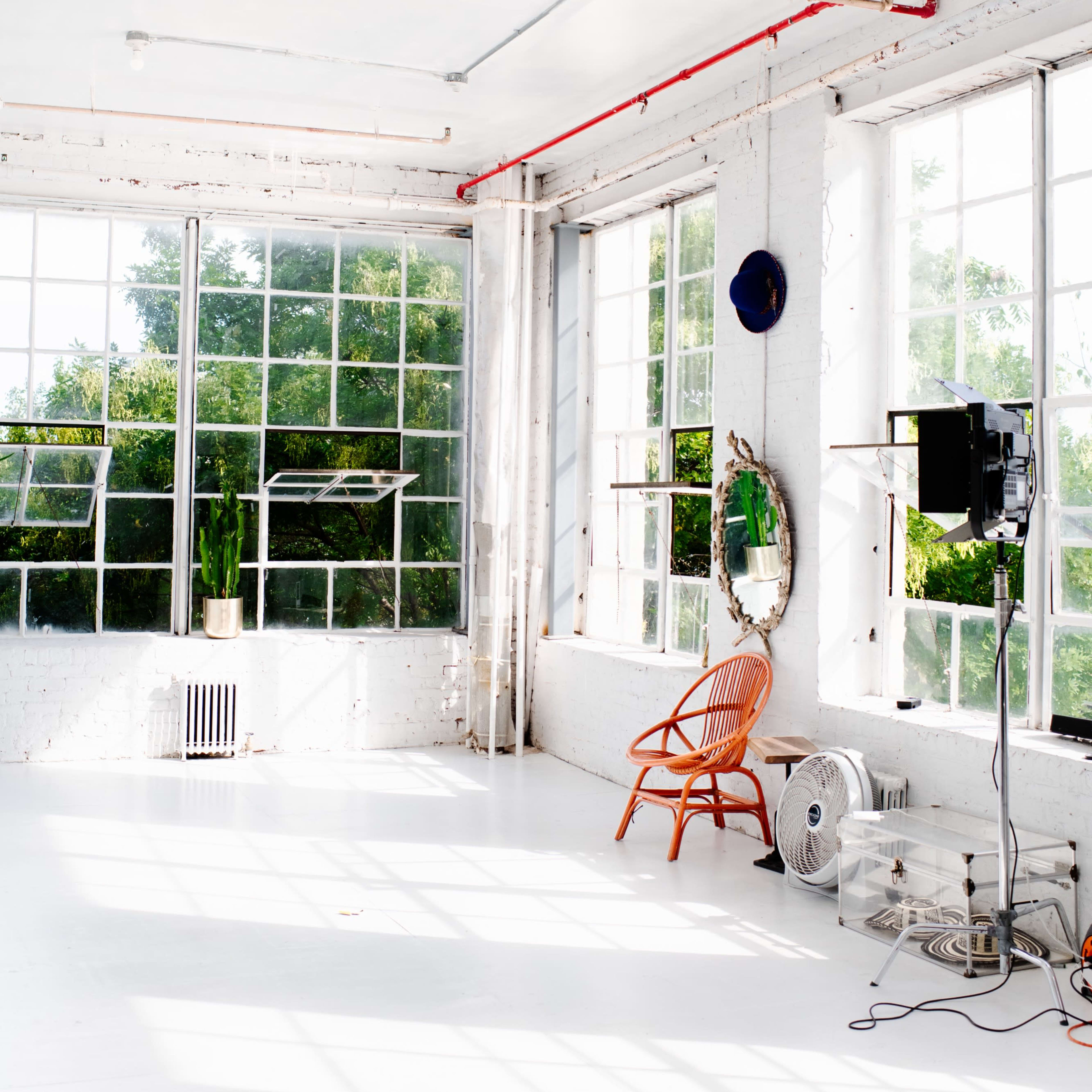 A spacious, well-lit studio with large windows, a single wooden chair, a fan, and a wall mirror.