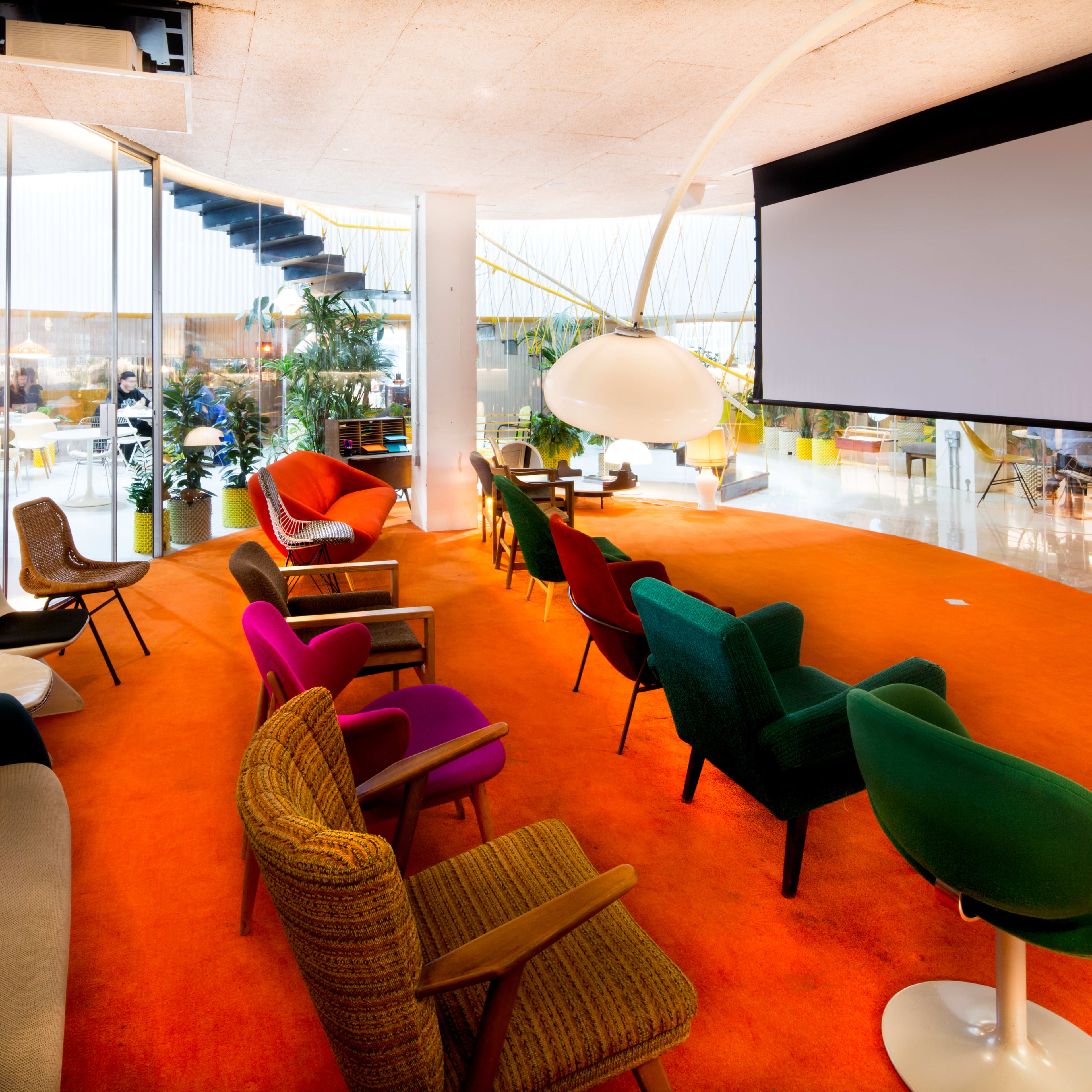 A lounge area features a variety of colorful chairs arranged around a large screen, with a vibrant orange carpet and plants in the background.