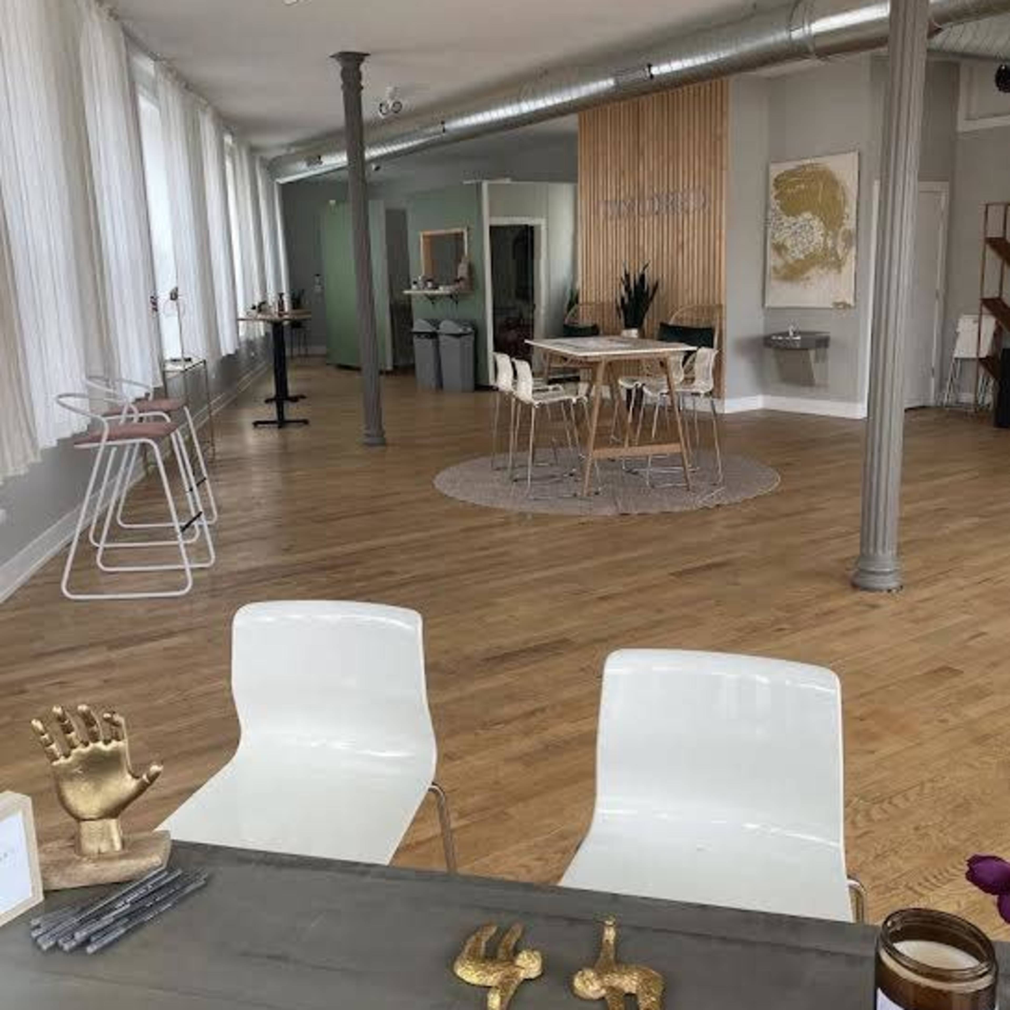 A spacious interior with wooden flooring, featuring a seating area with two white chairs, a table with decorative objects, and dining tables arranged in a well-lit space.