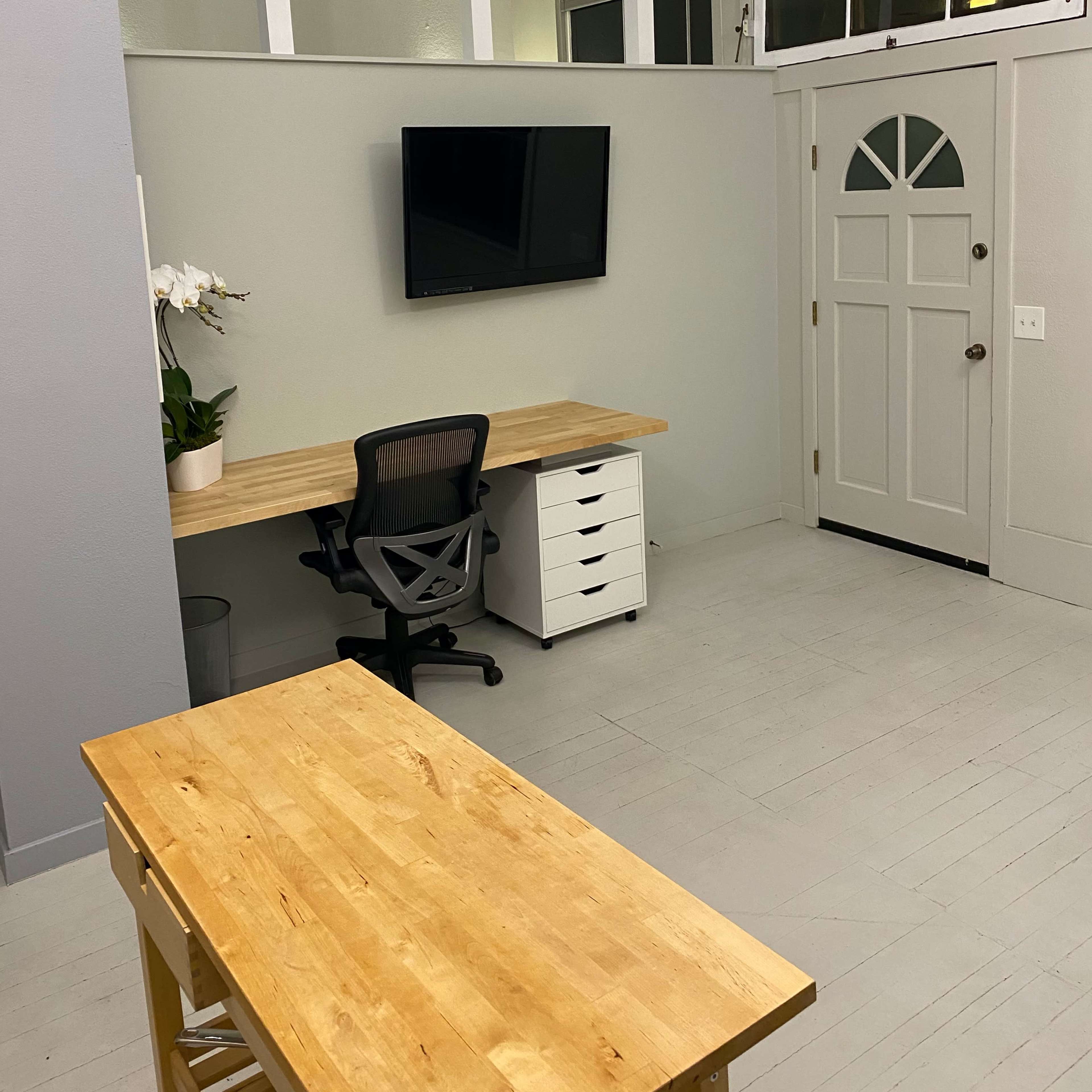 A minimalist home office features a wooden desk, white storage unit, and a wall-mounted TV, with a door leading outside visible in the background.
