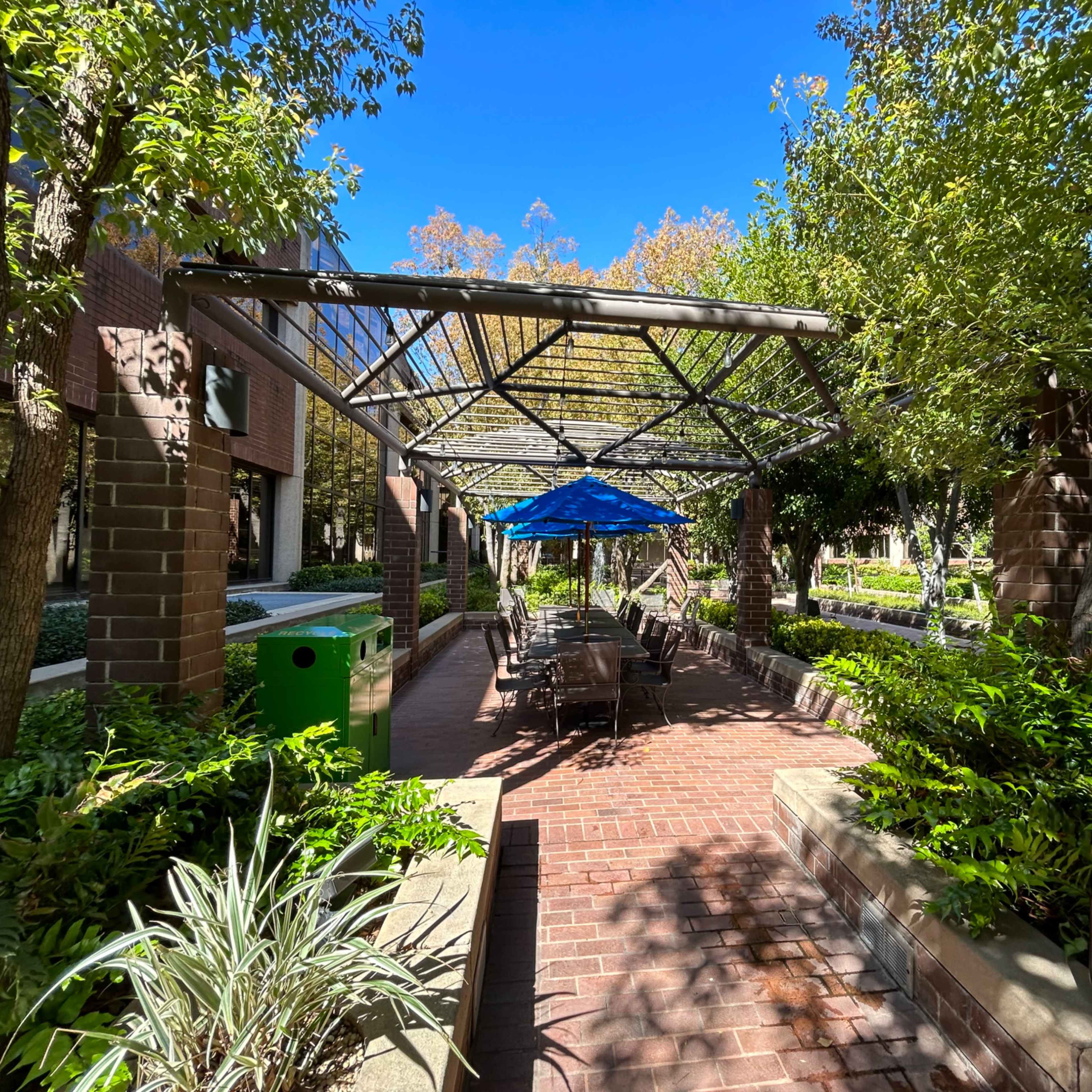 A covered outdoor seating area with blue umbrellas and brick pathways is surrounded by trees and greenery.
