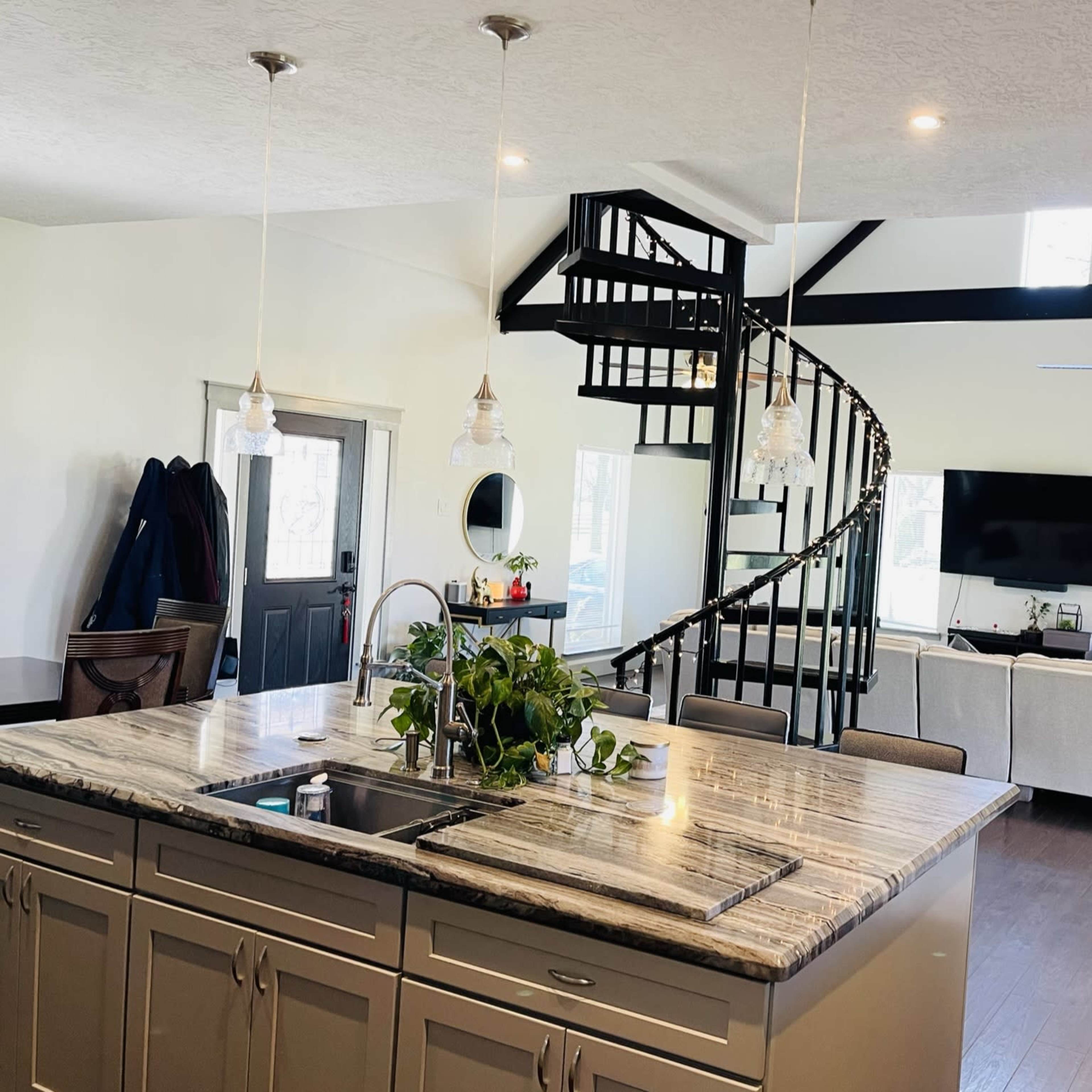 A modern kitchen with a large island, spiral staircase, and an open living area featuring a television and light-colored furniture.