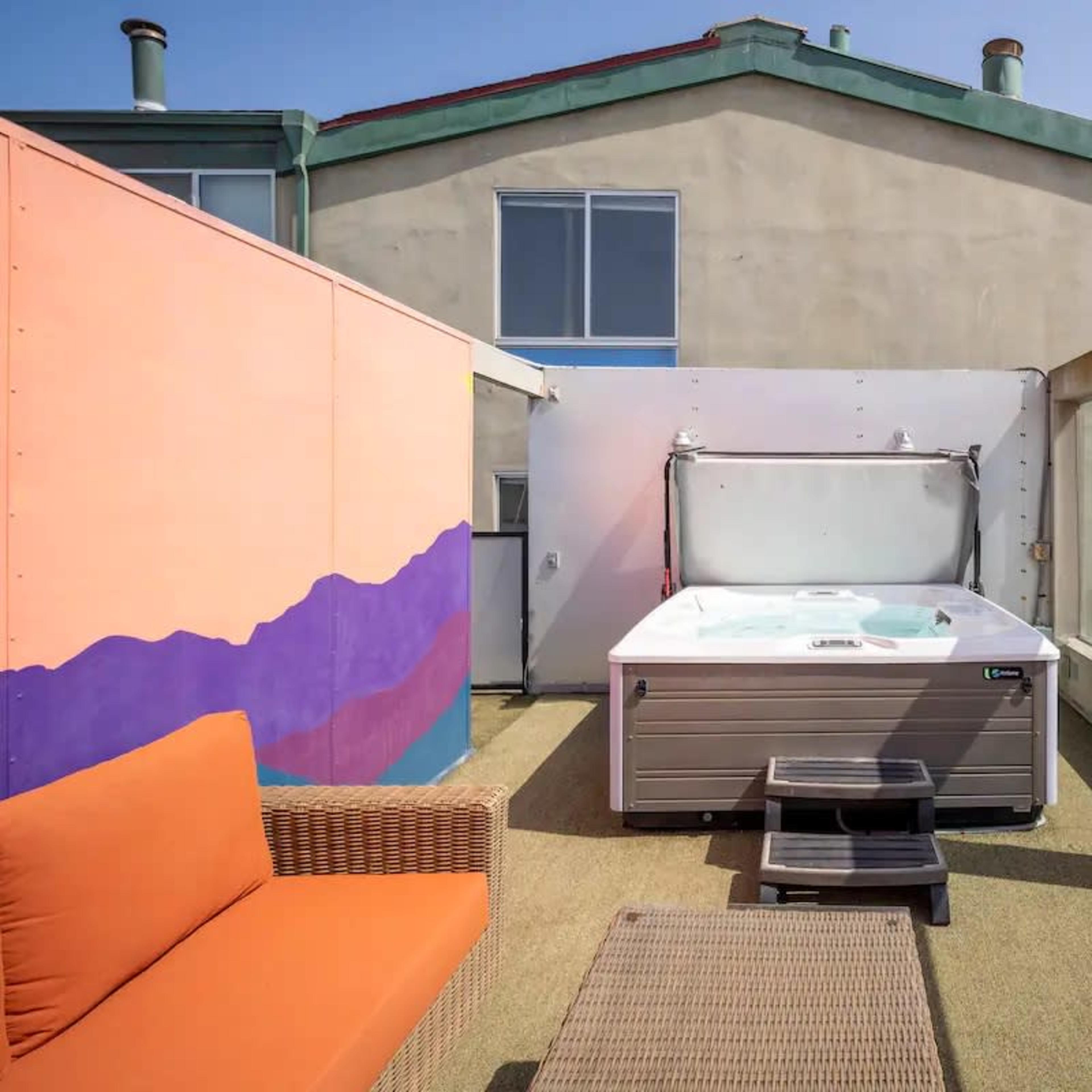 A hot tub sits on a deck surrounded by walls of glass, with a colorful mural and a view of the beach in the background.