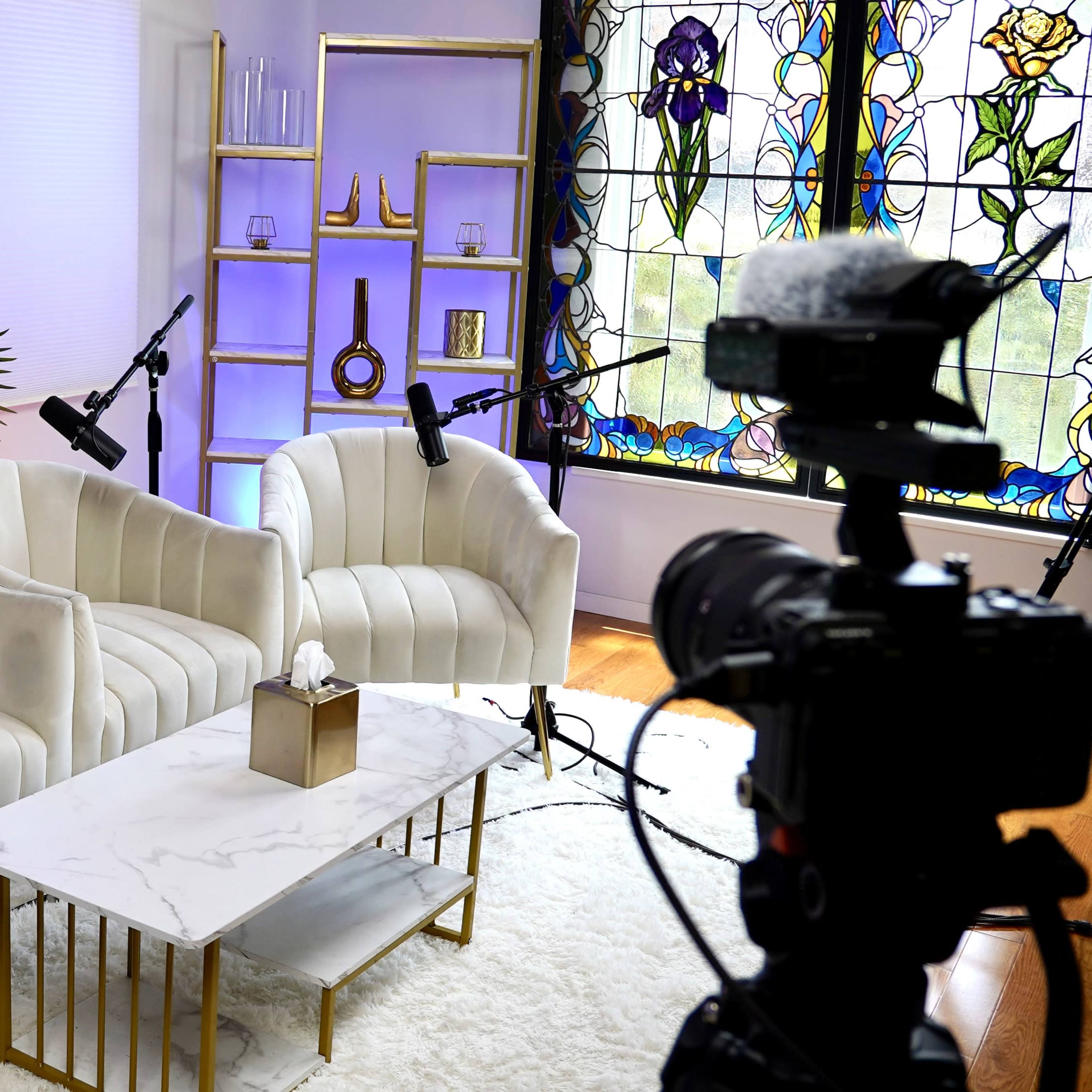 A modern studio setup with three white armchairs, a marble coffee table, and a stained glass window in the background.