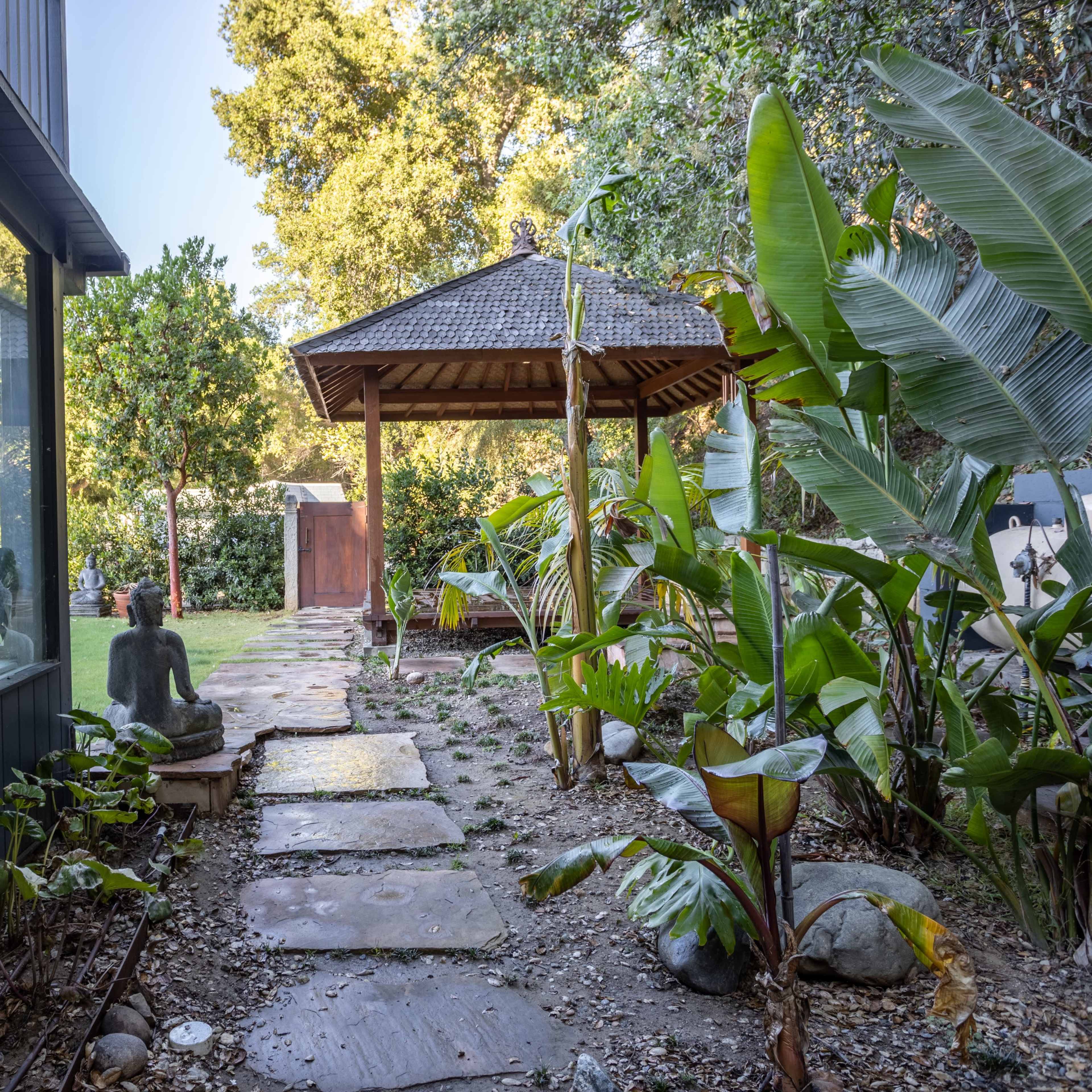 A stone path leads through a lush garden to a wooden gazebo surrounded by tropical plants and a statue.