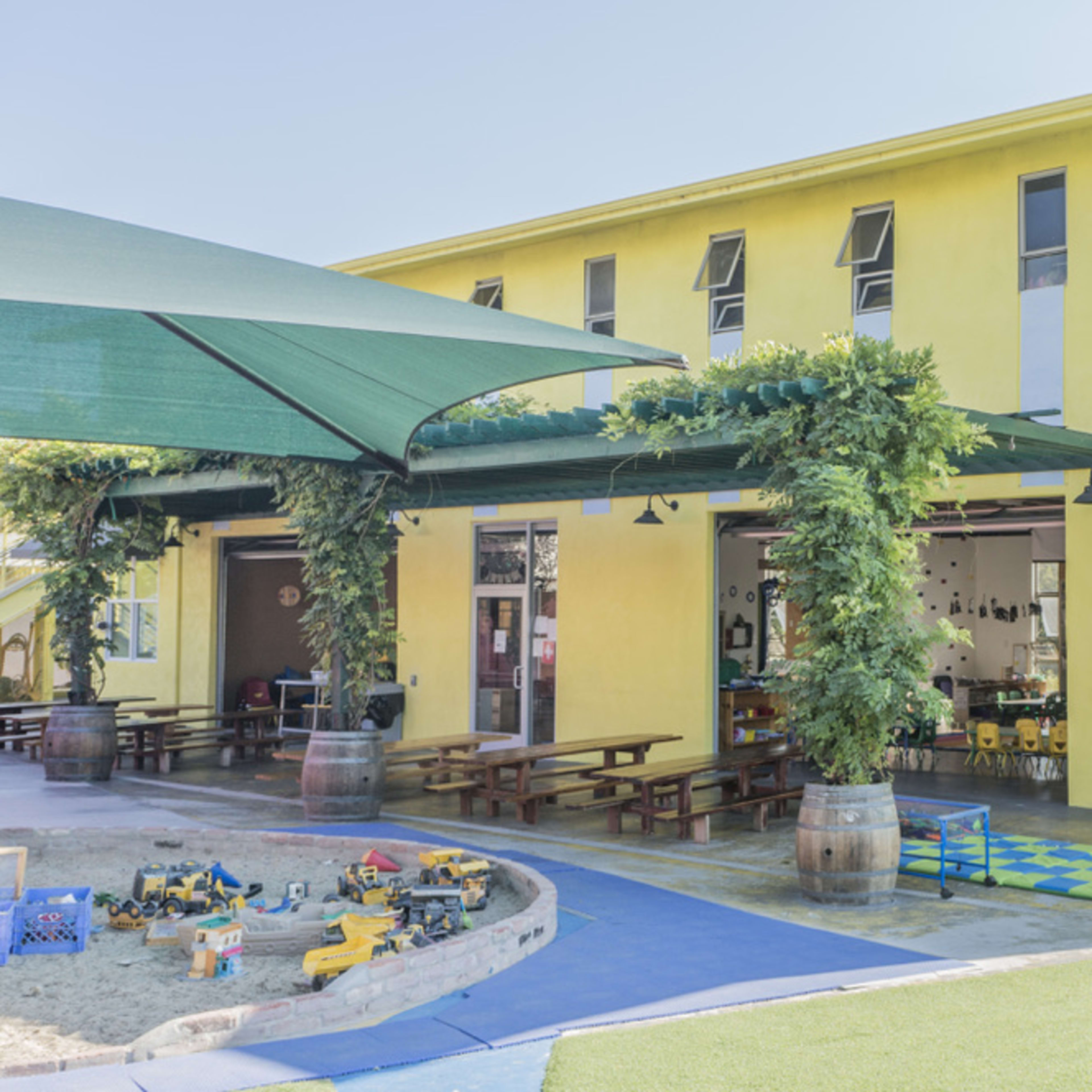 A yellow building with large windows houses a playground area featuring sand and colorful play equipment.