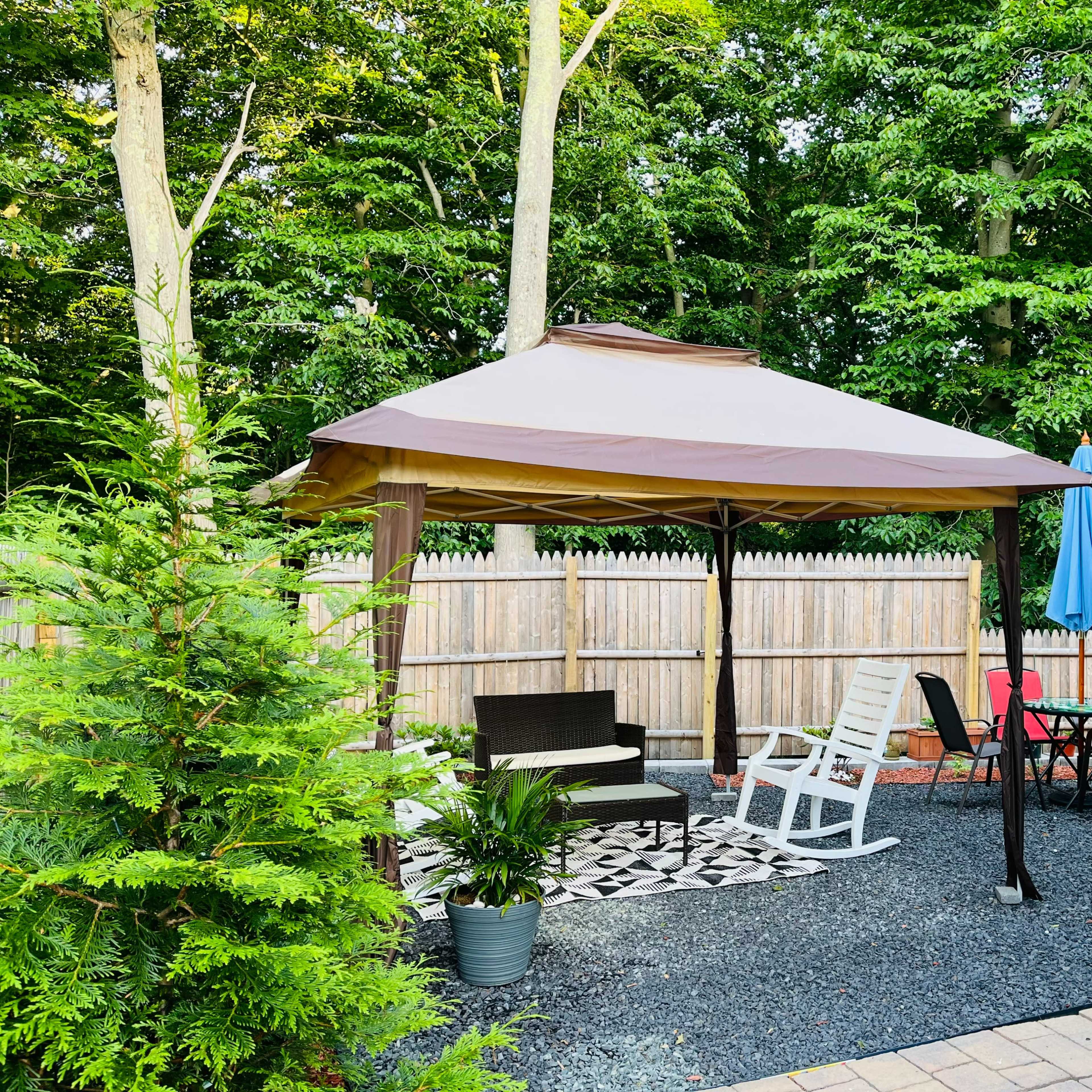 A gazebo with seating is set up in a landscaped backyard surrounded by trees and a wooden fence.