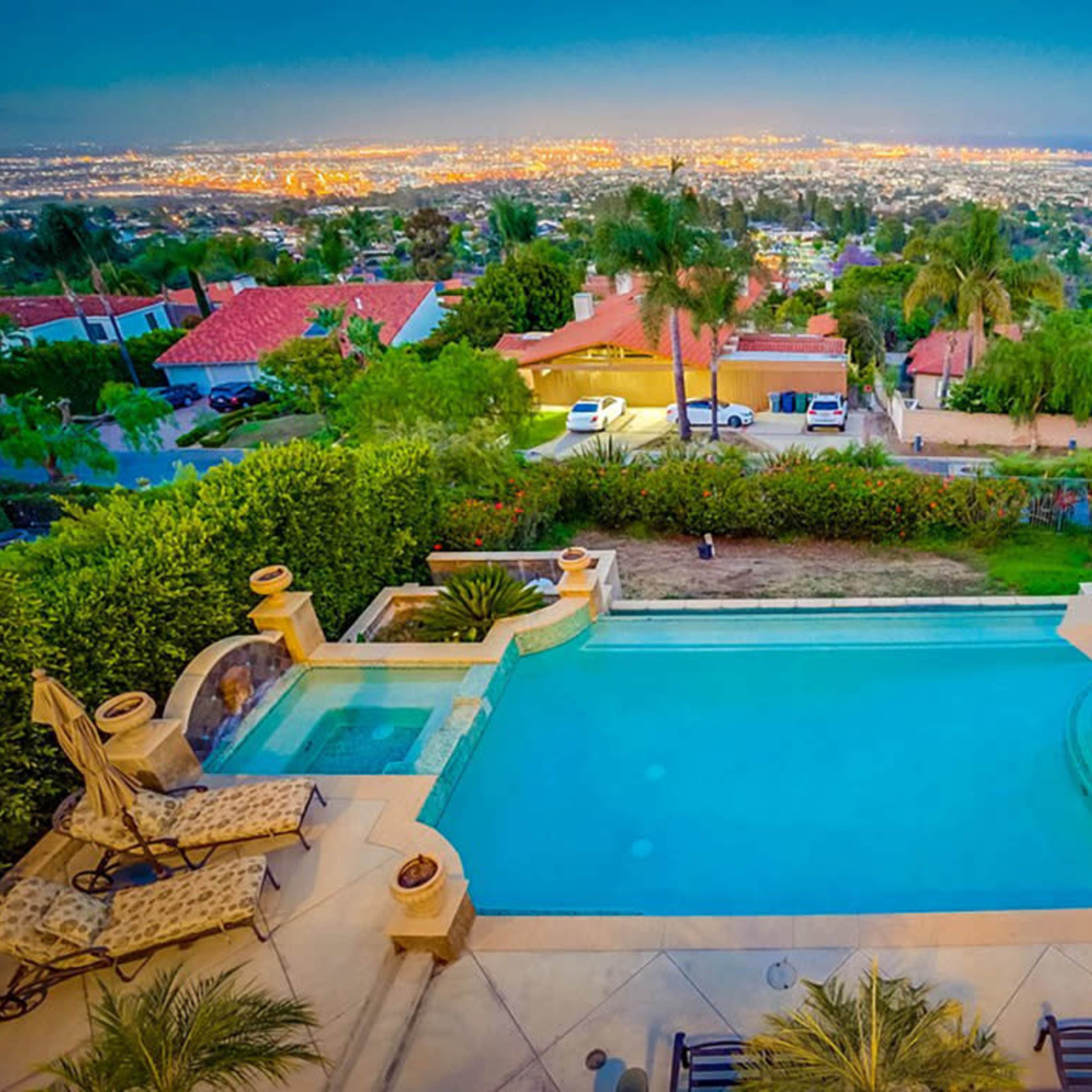 A backyard pool overlooking a city skyline illuminated at dusk, surrounded by lush greenery and lounge chairs.