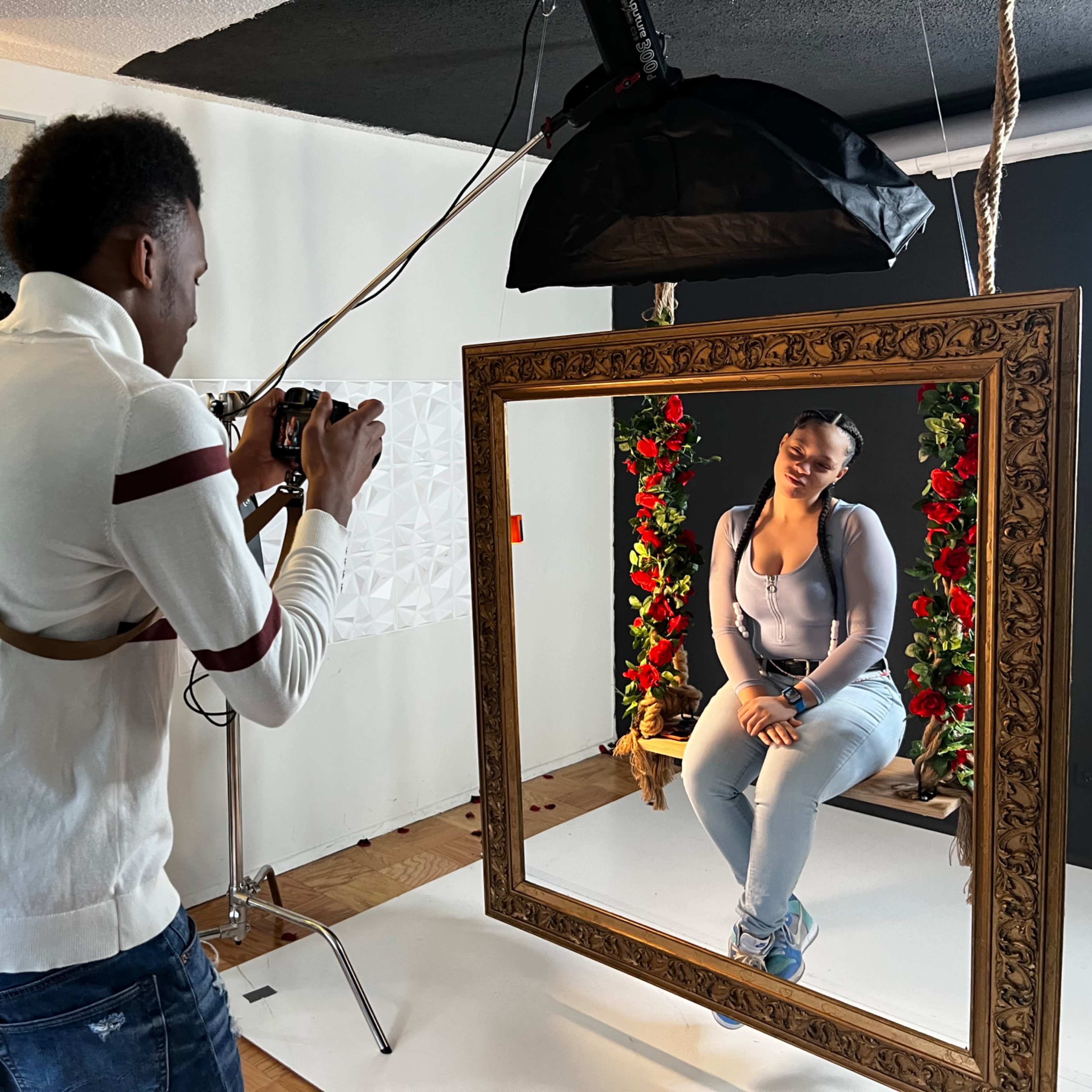 A photographer captures a portrait of a model seated in a decorative frame surrounded by floral arrangements in a studio setting.