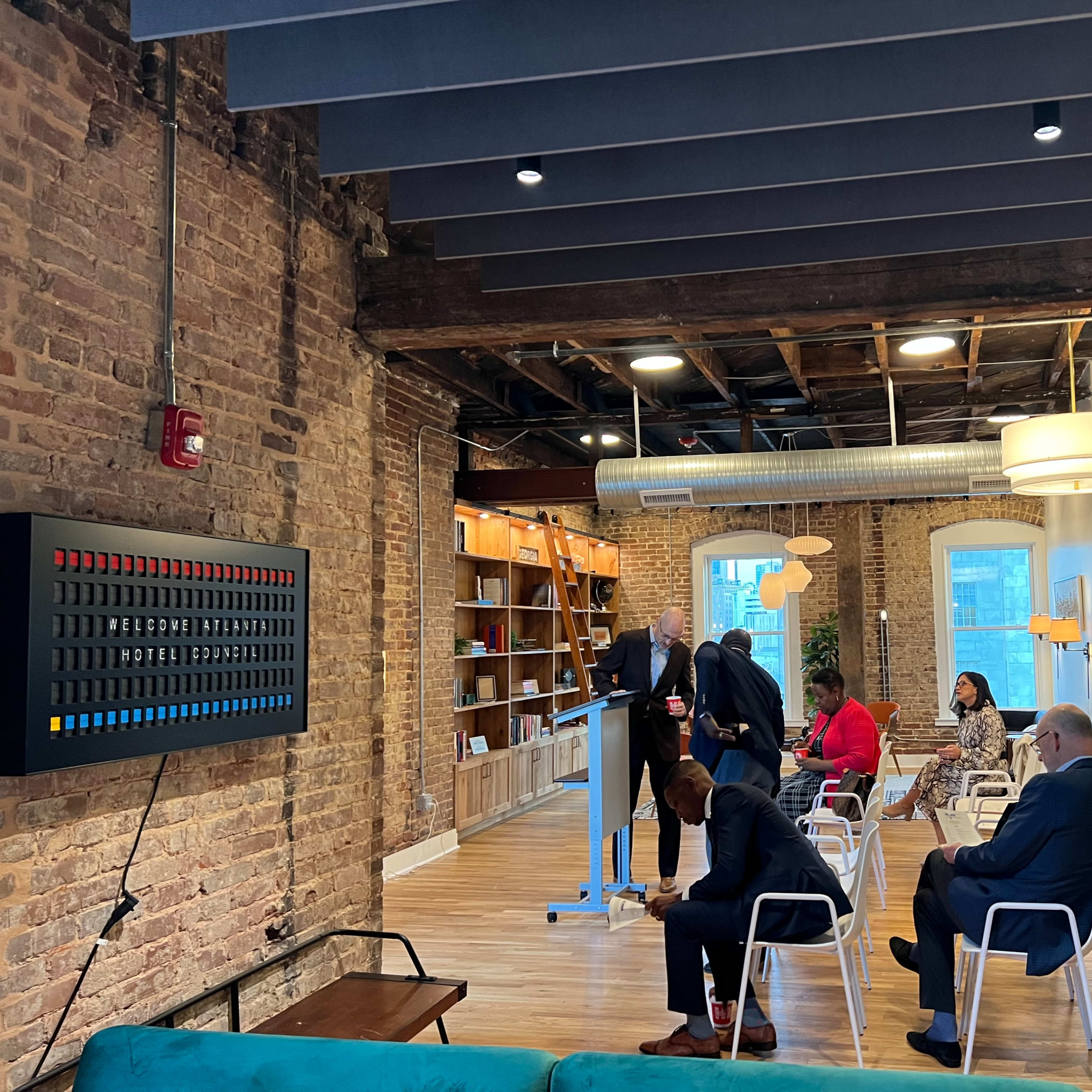 A group of people are gathered in a modern office space with exposed brick walls, some standing near a presentation setup while others are seated.