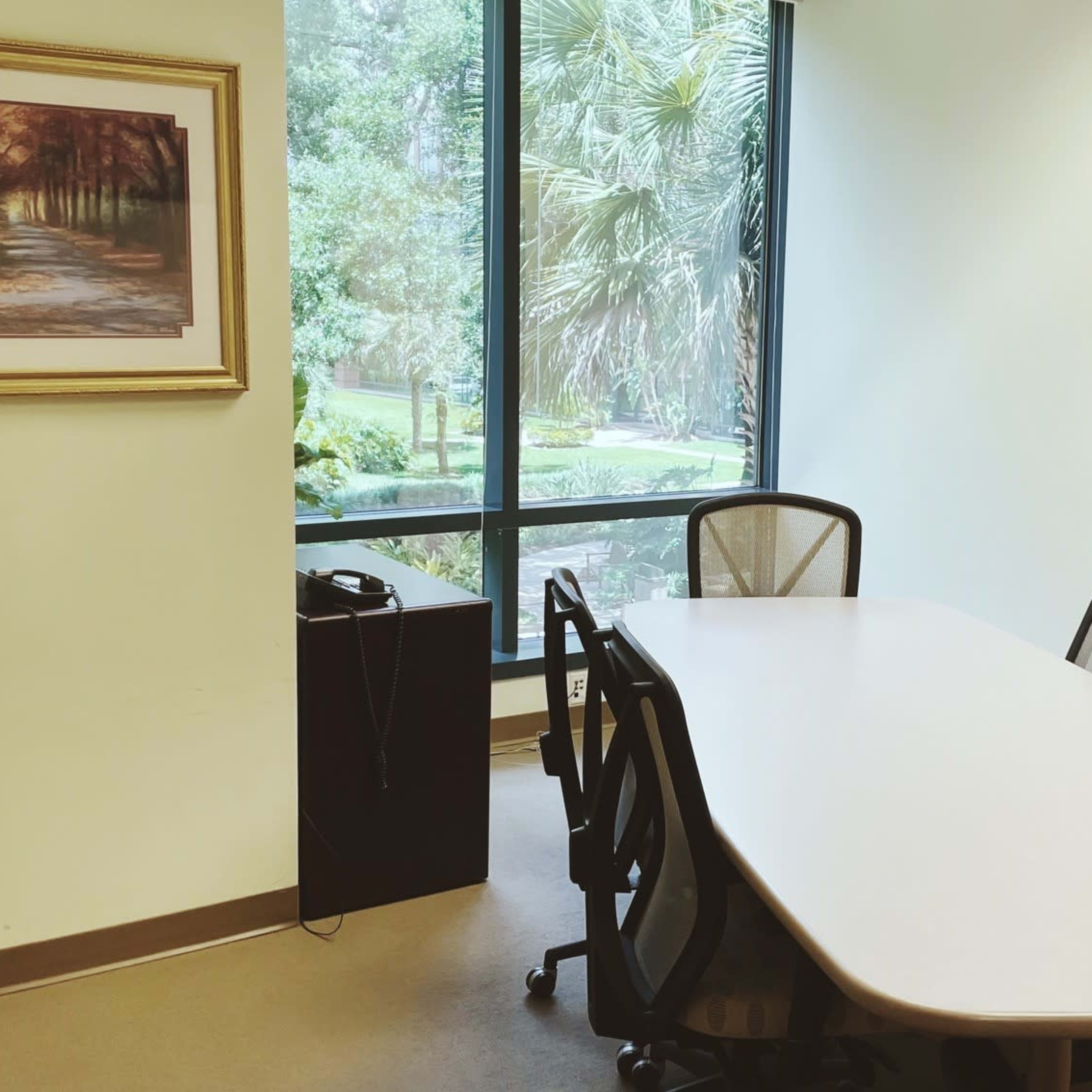 A conference room features a long table surrounded by chairs, a whiteboard, a framed landscape painting, and a window with a view of greenery.