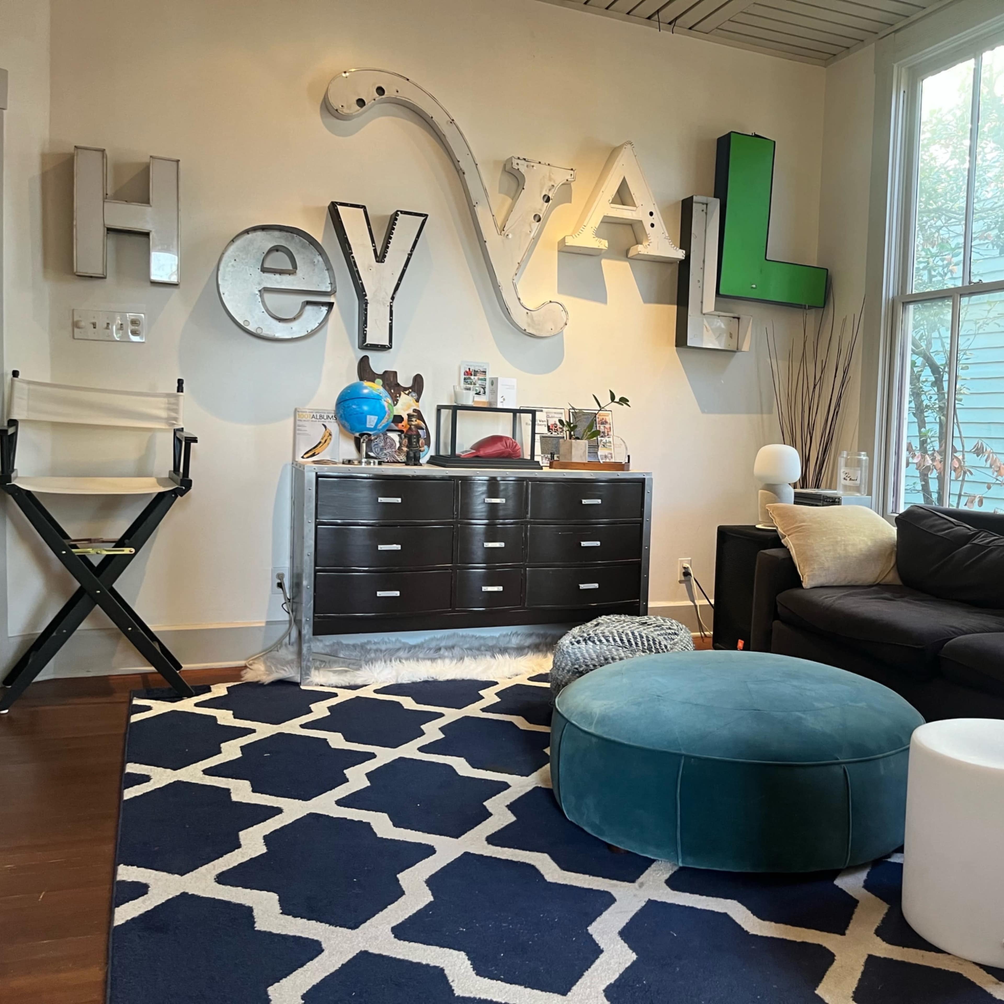The scene features a living room with a dark sofa, a blue pouf, a white side table, and large decorative letters spelling "Hey Y'all" on the wall.