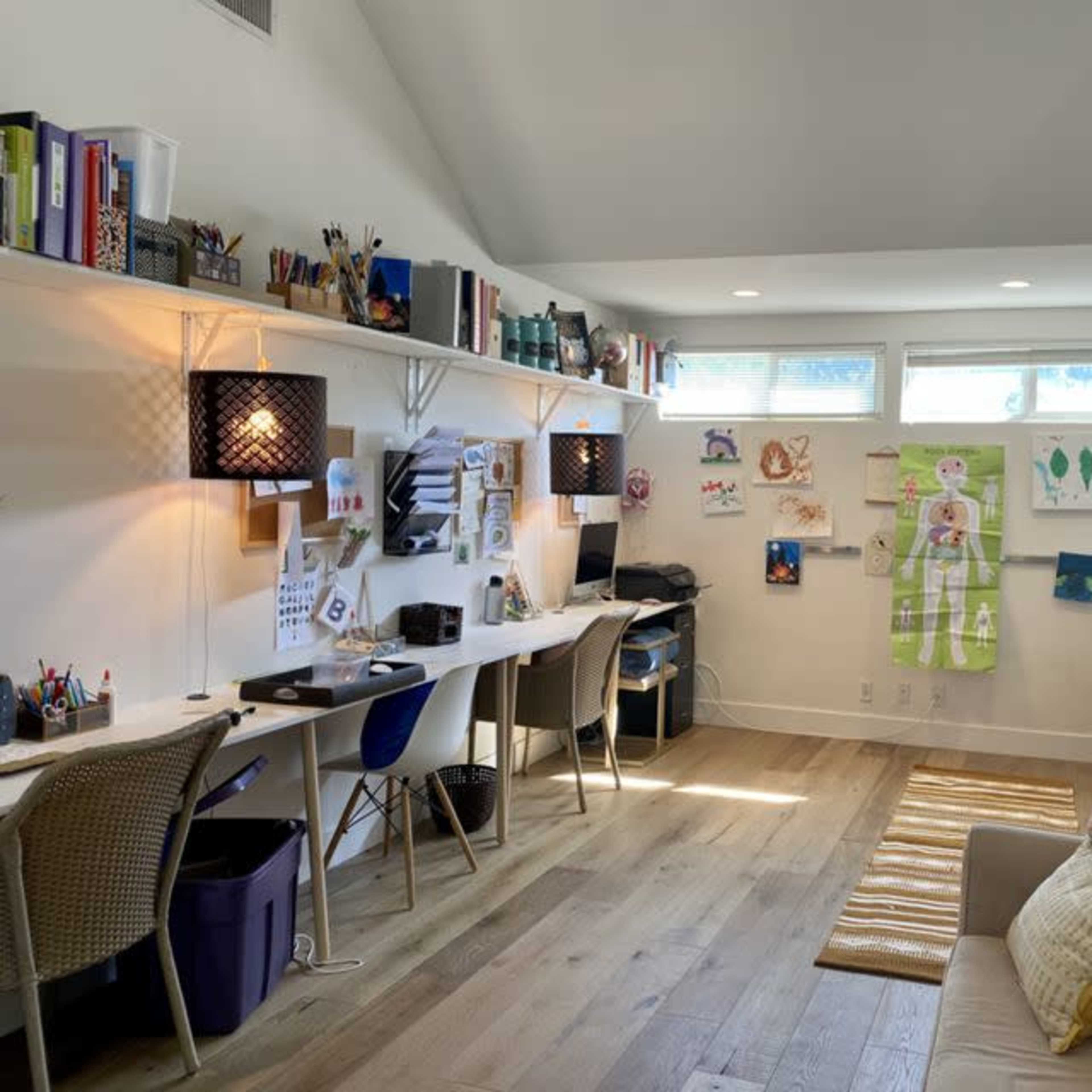 A spacious room with two work desks, shelves filled with art supplies and books, and a couch, all illuminated by natural light from large windows.