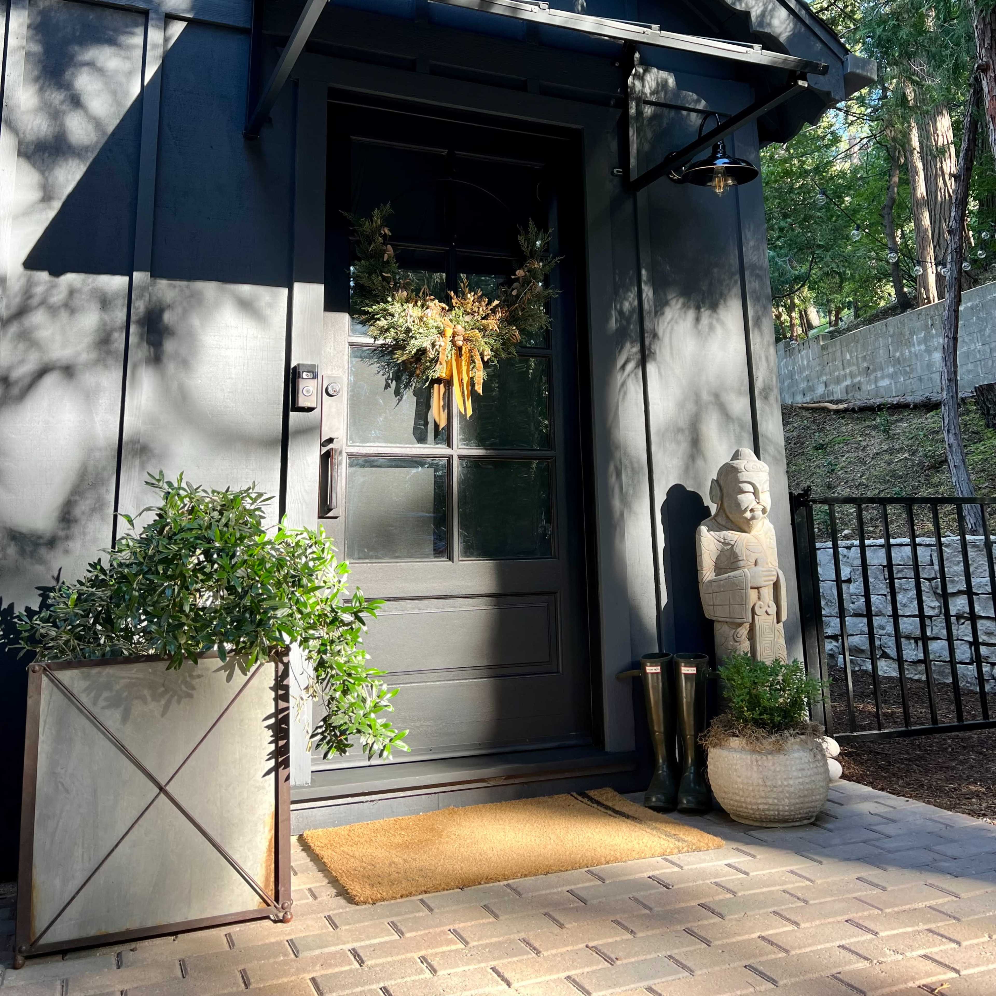 A dark exterior door adorned with a seasonal wreath, flanked by potted plants and decorative items, set against a backdrop of greenery.