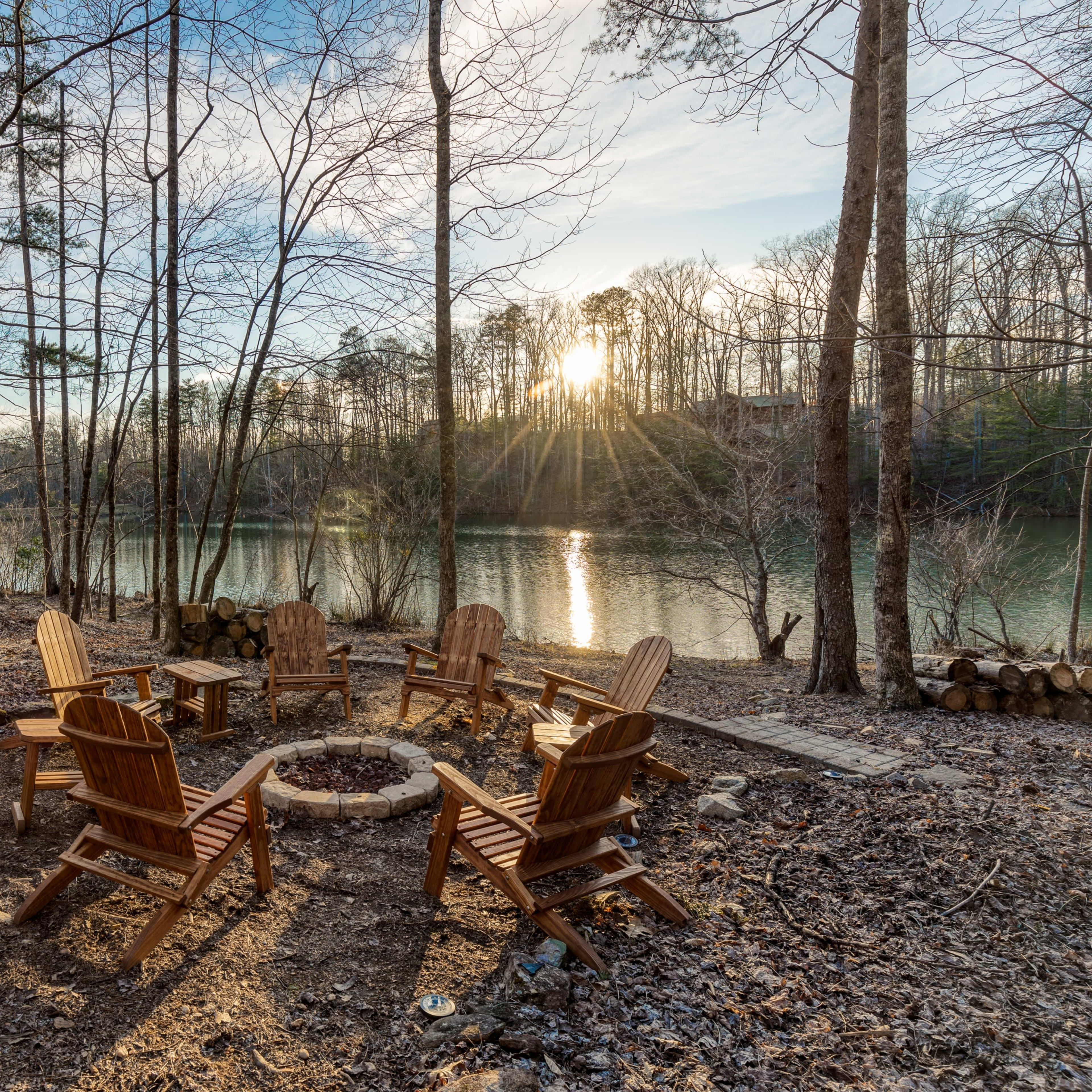 A circle of wooden chairs surrounds a fire pit by a lake, framed by trees reflecting the setting sun.
