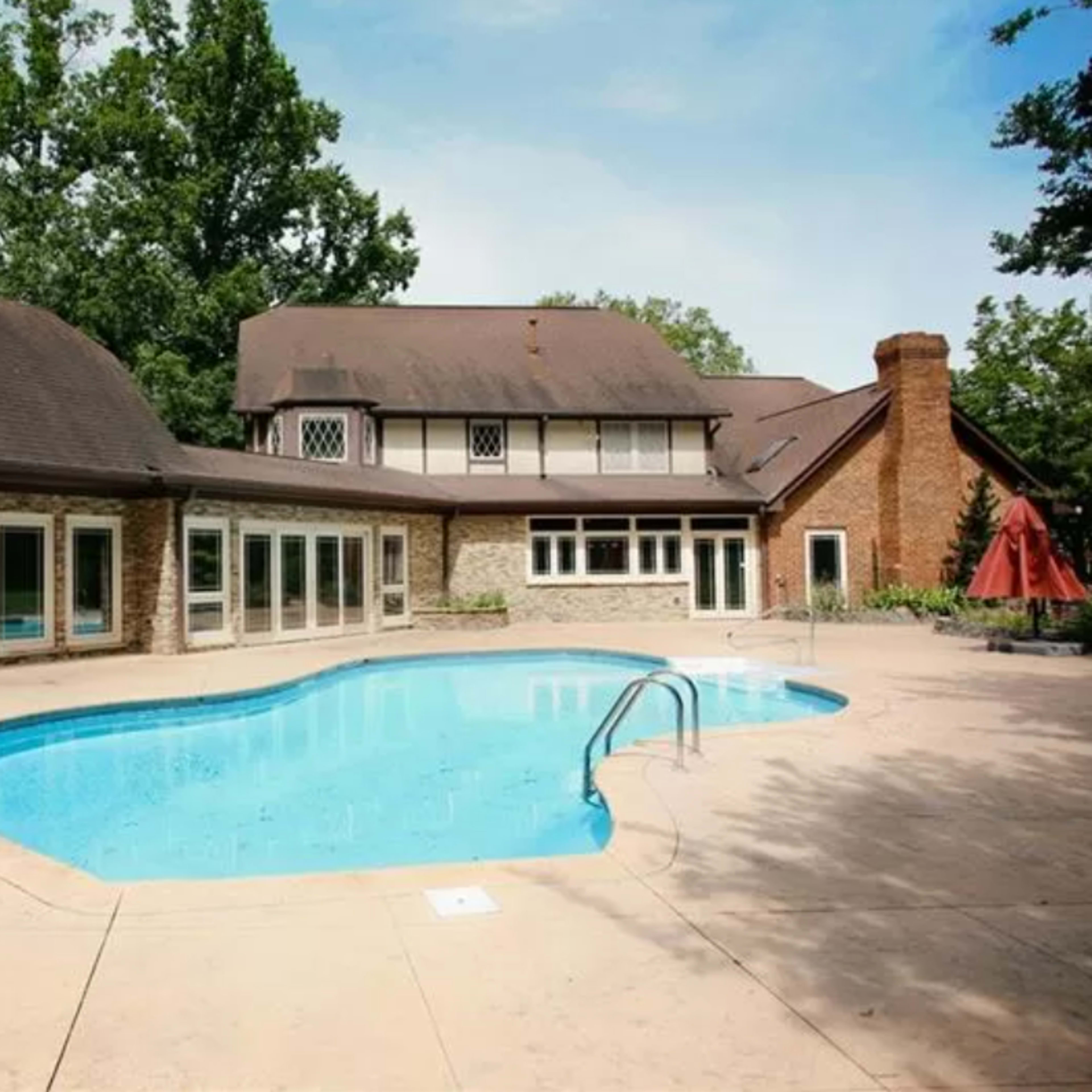 A large, rectangular swimming pool is surrounded by a stone and tile patio, with a two-story house featuring a mix of brick and timber architecture in the background.