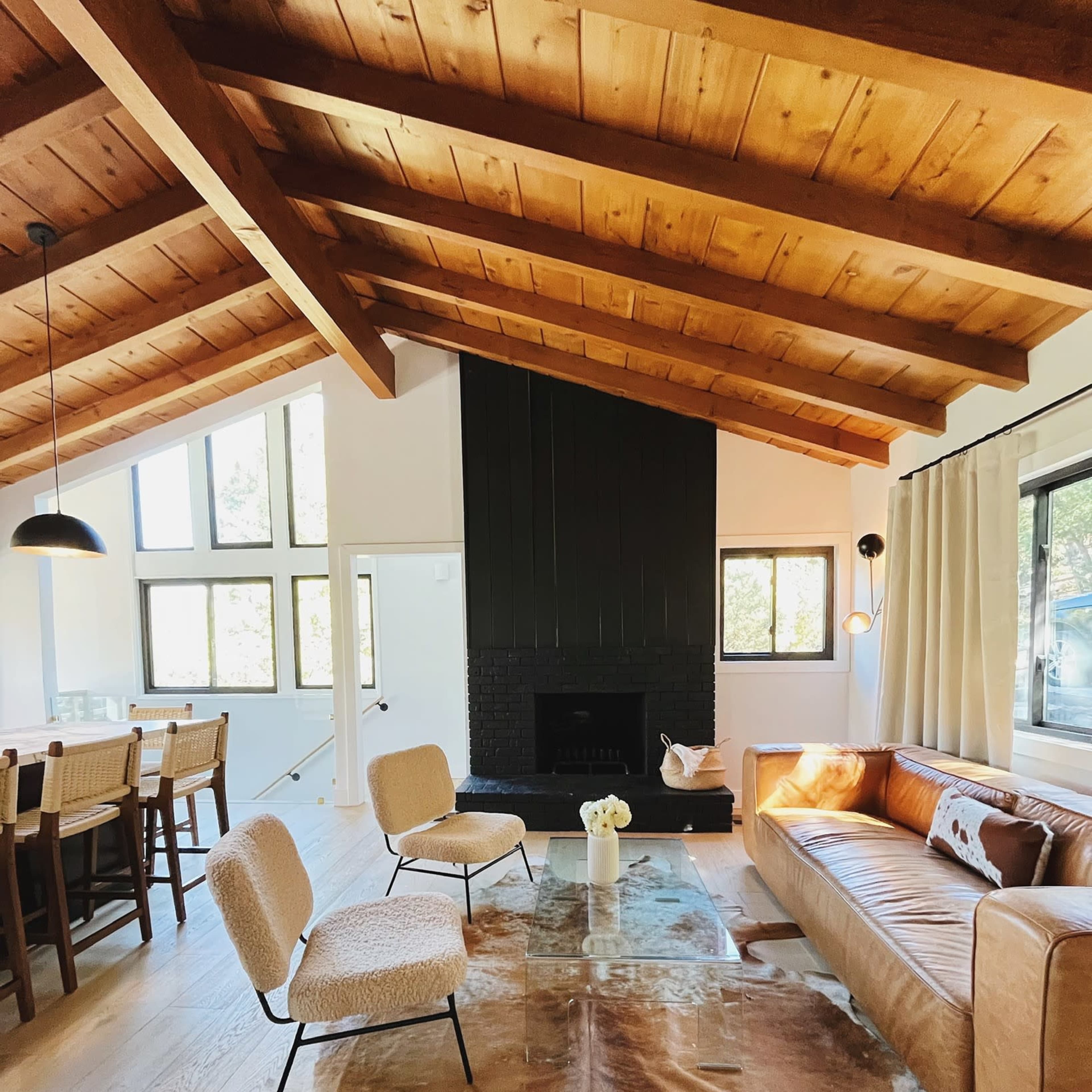 The living space features a brown leather sofa, two upholstered chairs, a rustic cowhide rug, and a fireplace, with large windows allowing natural light to fill the room.