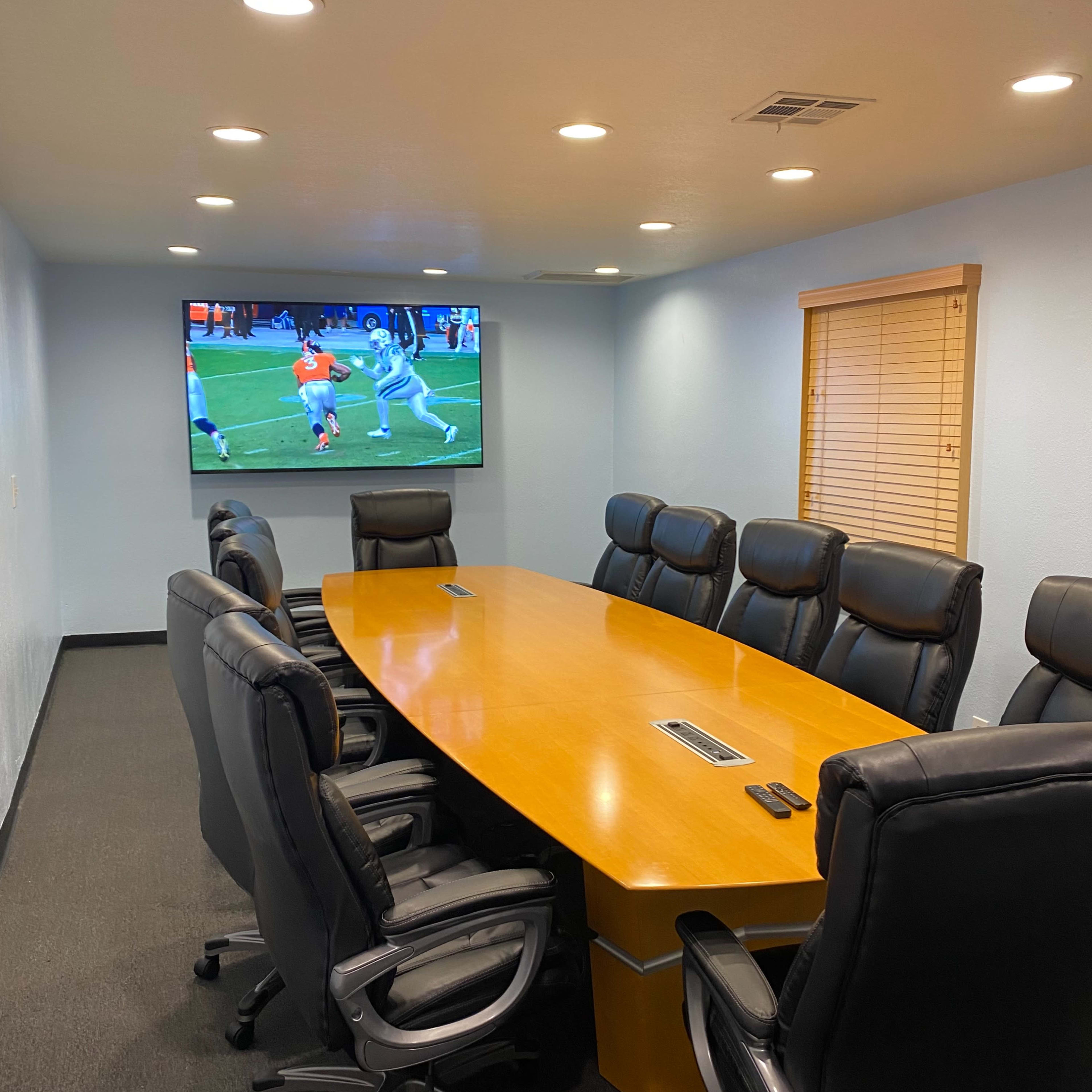 A conference room features a long wooden table surrounded by black leather chairs, with a television displaying a sports event on the wall.
