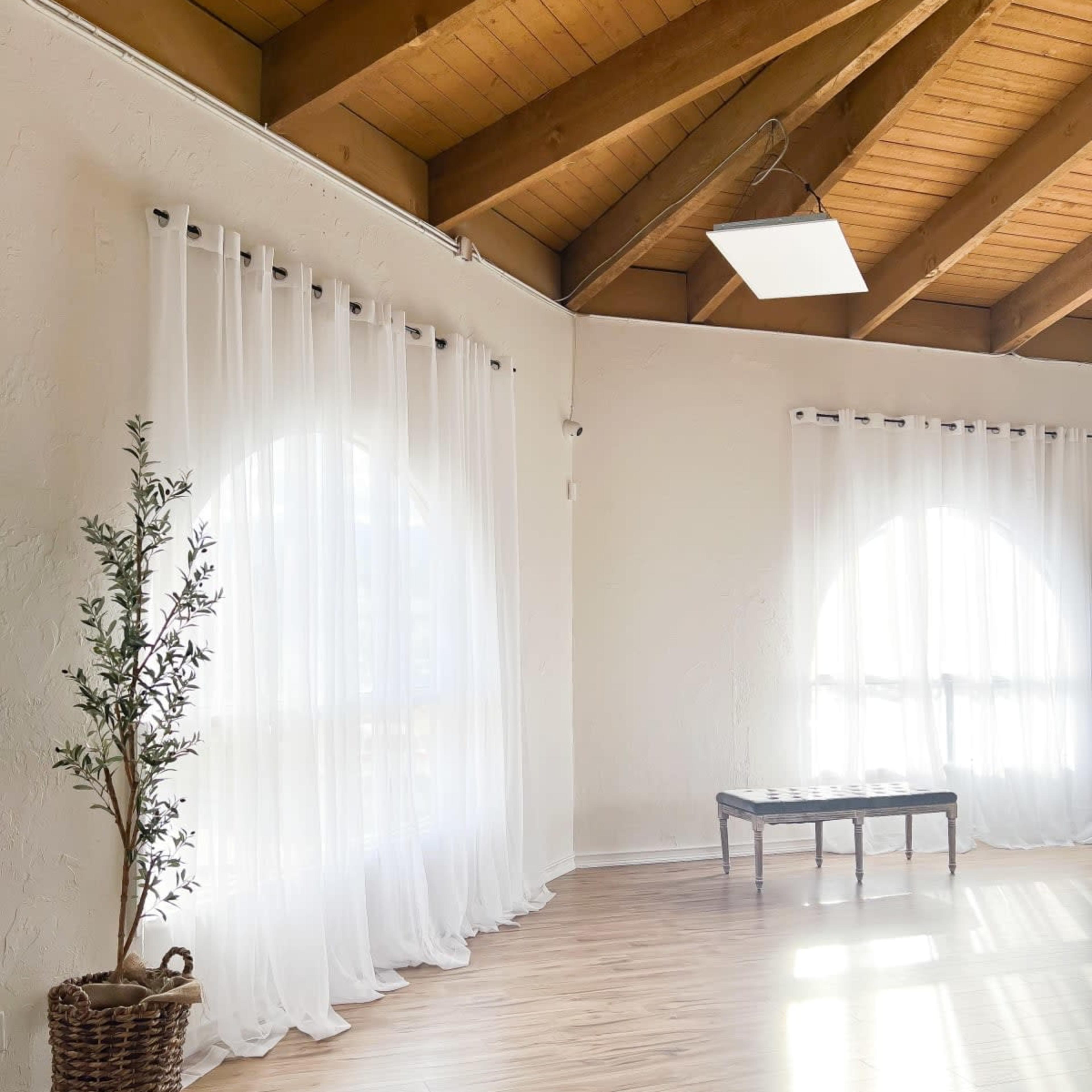 A spacious interior features large windows with sheer curtains, wooden beams, and a simple bench beside a potted plant.